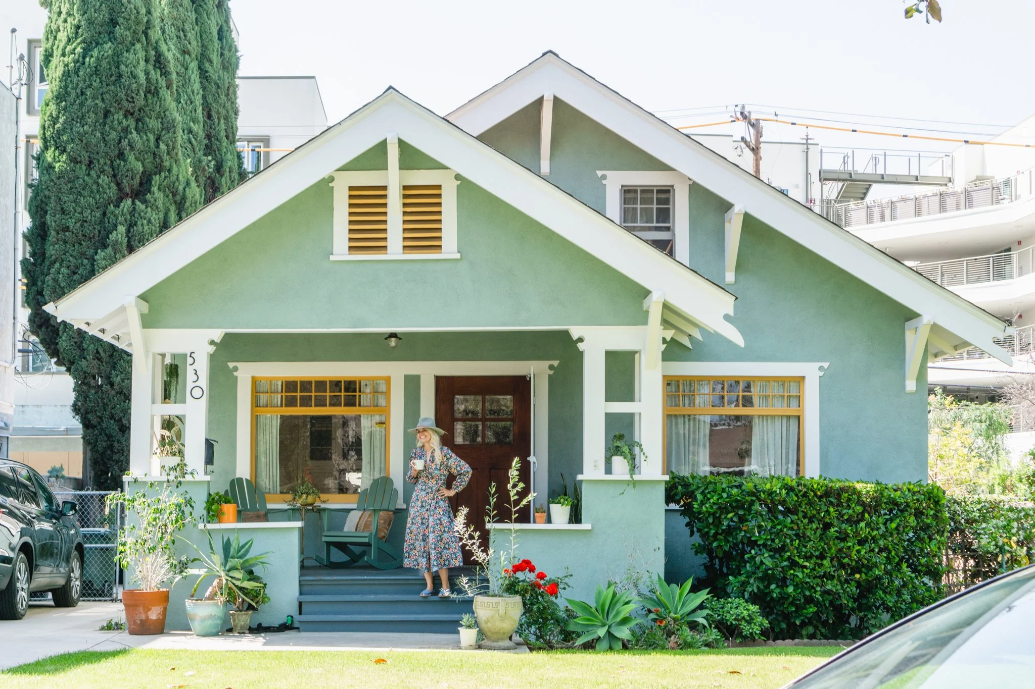 A woman in a floral dress and hat standing on the front steps of a green house, holding a cup, with potted plants and outdoor furniture in the yard.