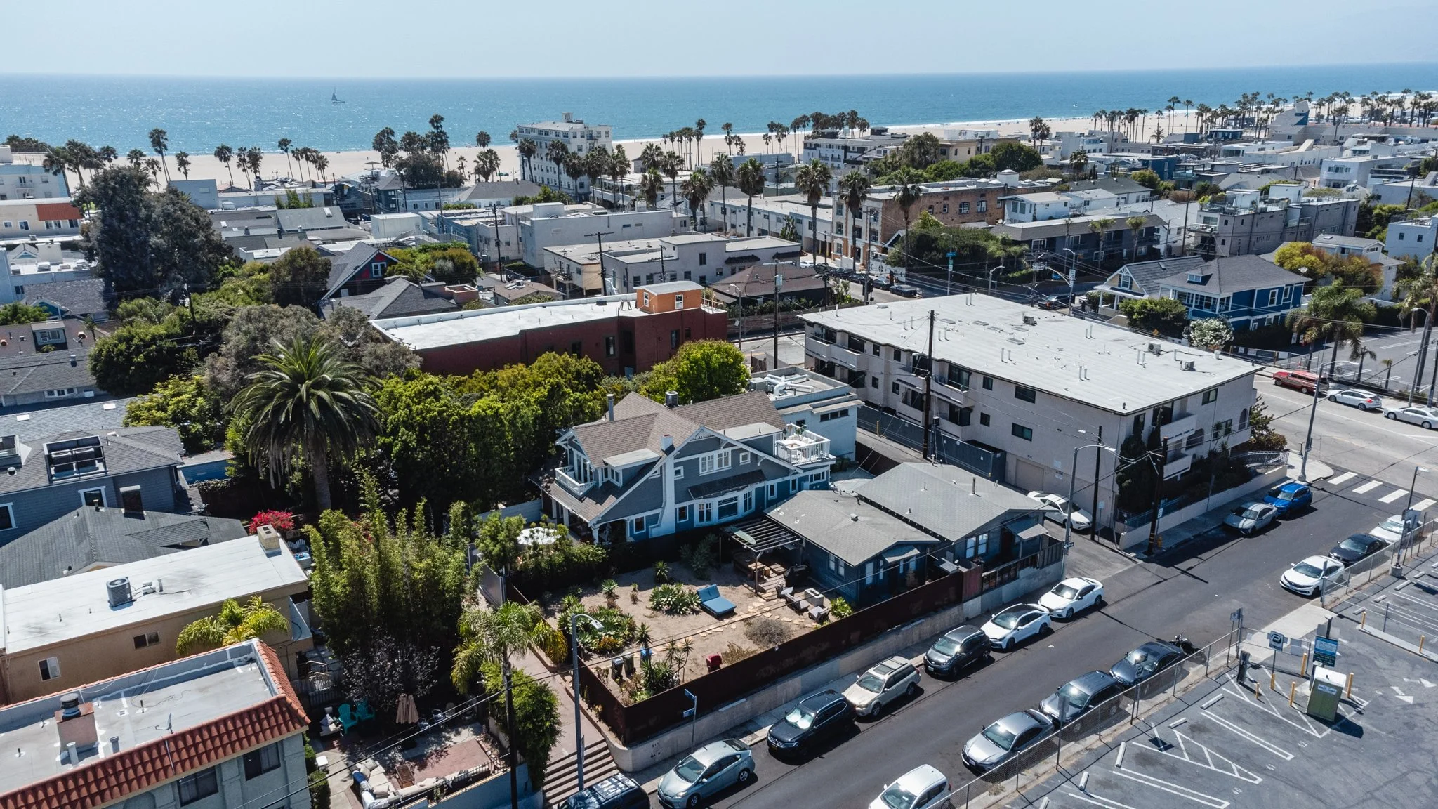 Aerial view of coastal neighborhood with houses, palm trees, and a beach with ocean in the background, sunny day.