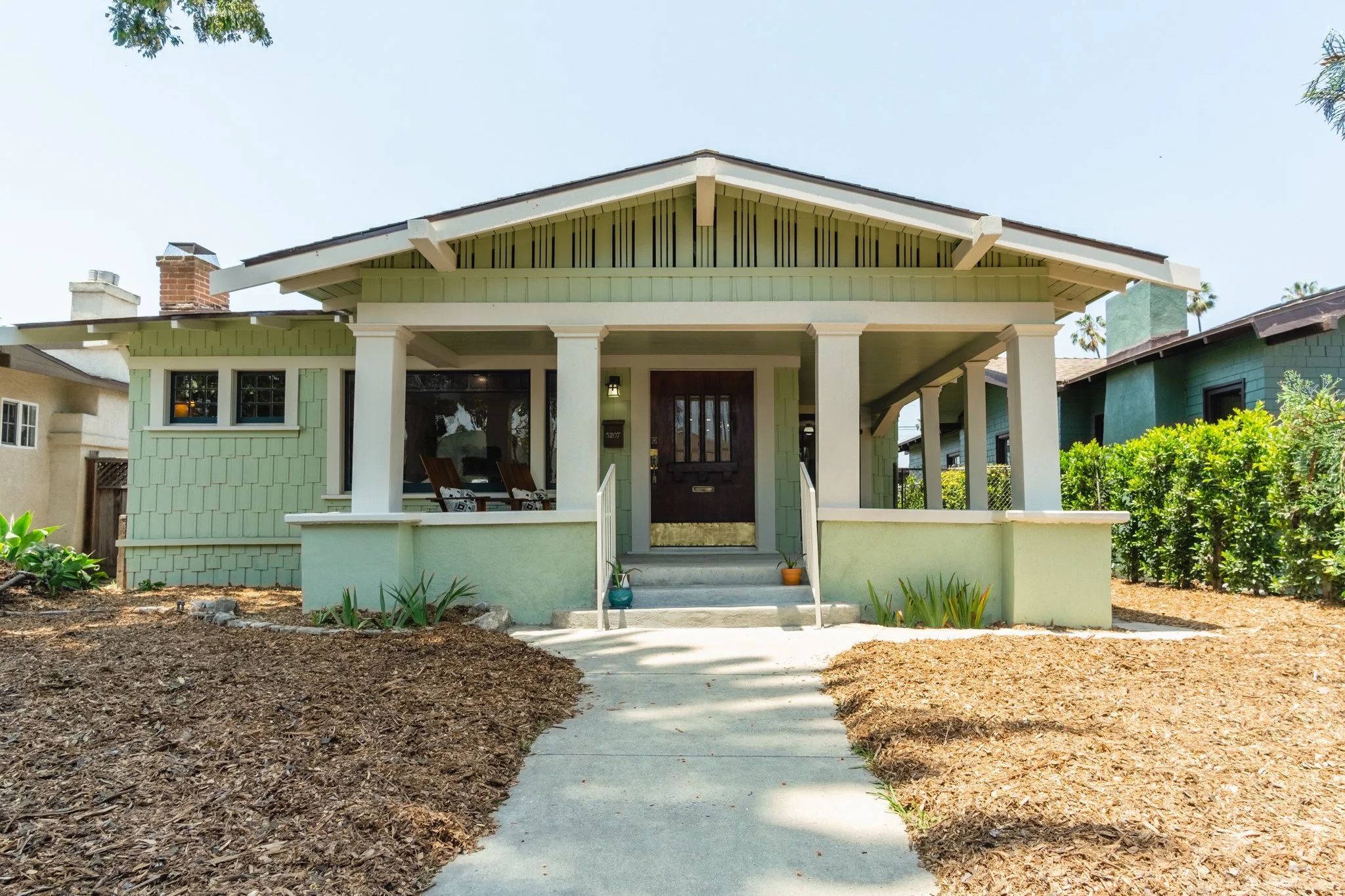 Front view of a light green house with a covered porch, a wooden front door, and stairs leading up to the entrance, surrounded by a garden with plants and a sidewalk.