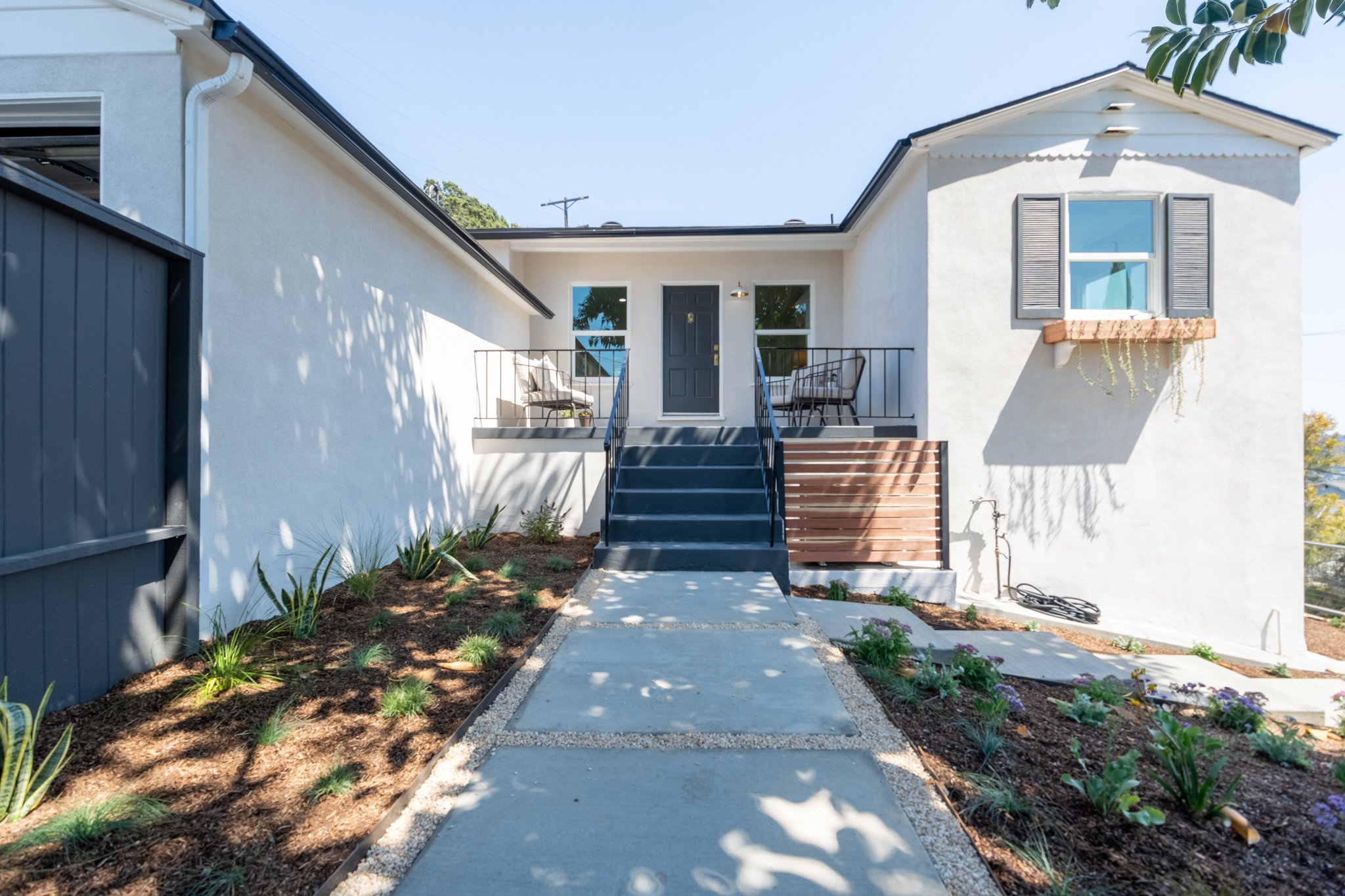 A white residential house with a small front porch, two chairs, and a door, seen from the sidewalk with a small garden and plants along the pathway.