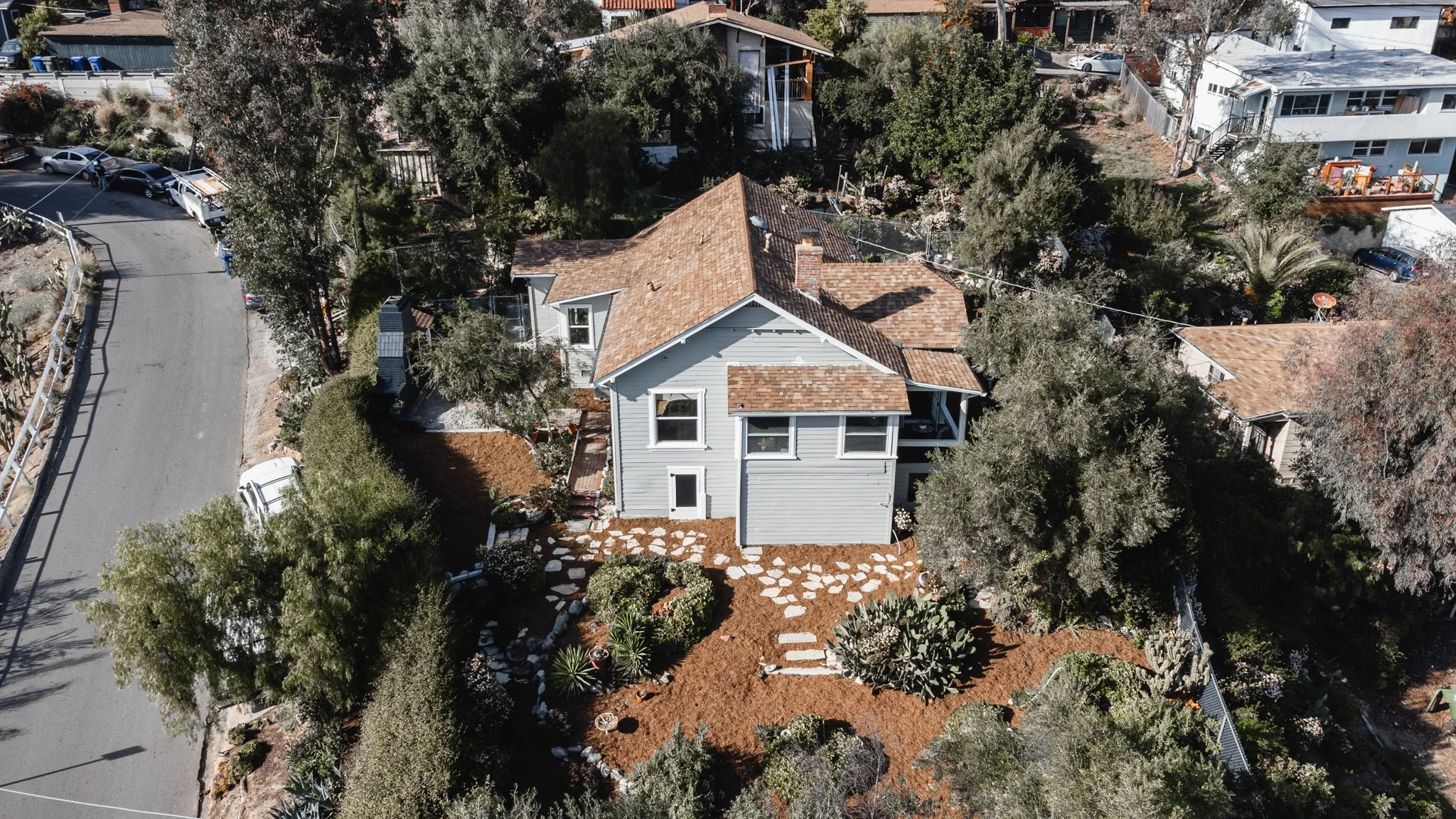 An aerial view of a house with a brown shingle roof surrounded by green trees and a landscaped yard with stepping stones, in a neighborhood with other houses and parked cars.