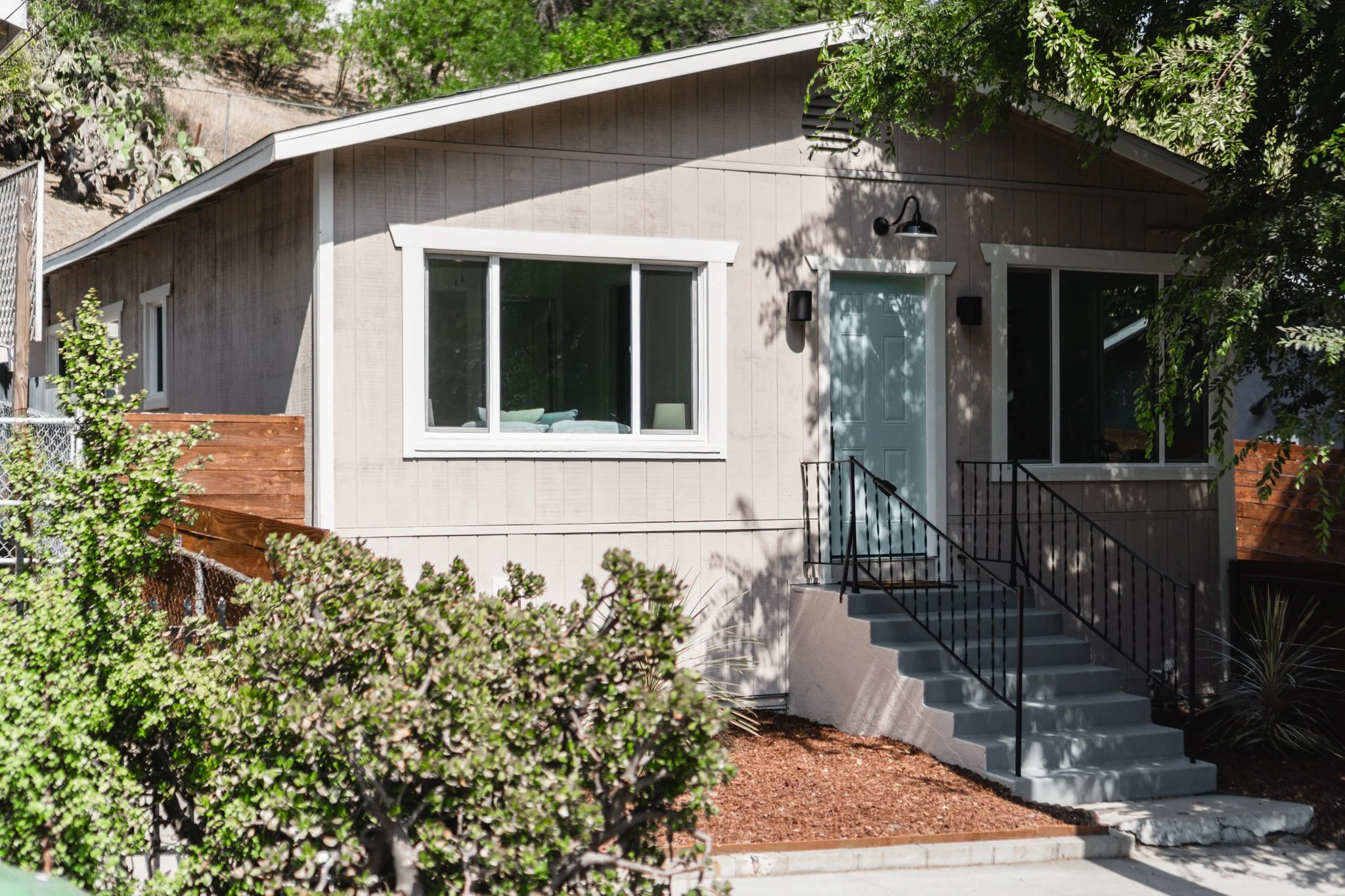 A light-colored house with a stepped front porch and black railing, surrounded by trees and bushes.