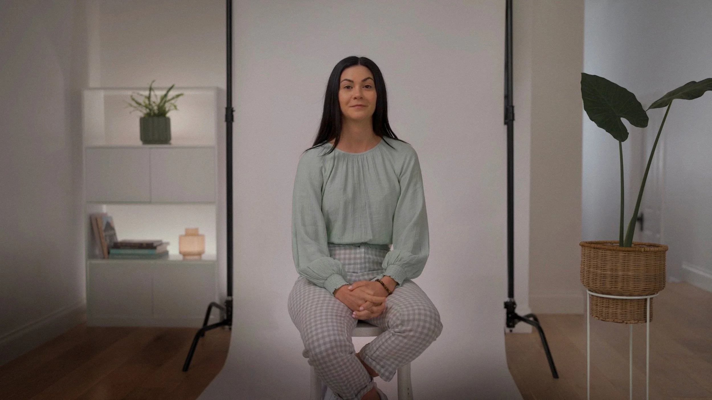 Woman sitting on a stool in front of a white backdrop with a potted plant beside her.