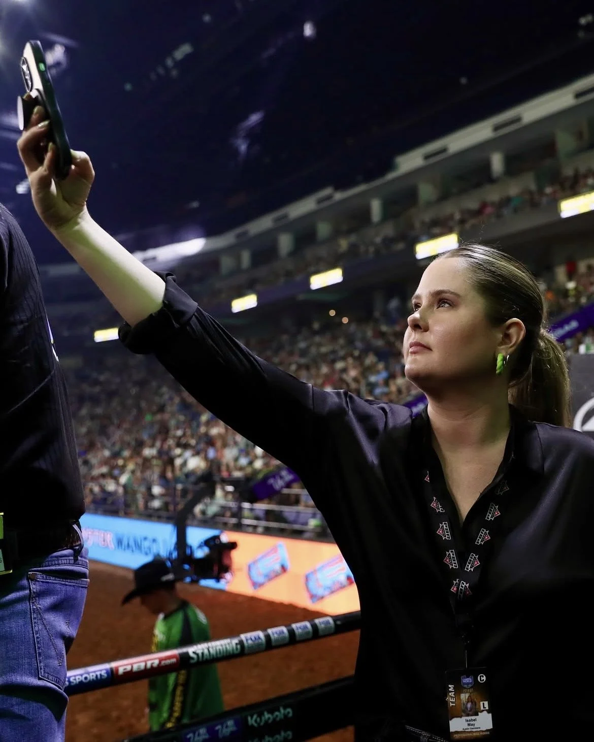 A woman with a ponytail and earrings filming on her phone at a large sports stadium filled with spectators.
