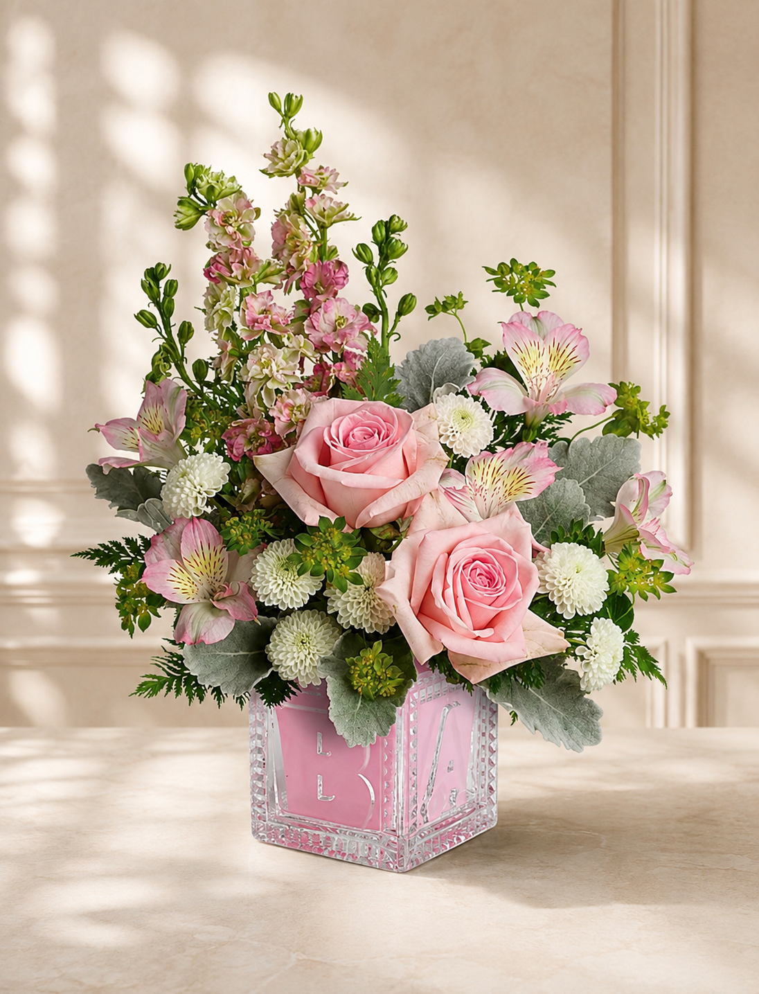 A floral arrangement with pink roses, pink and white alstroemeria, white chrysanthemums, and greenery in a pink decorative vase.