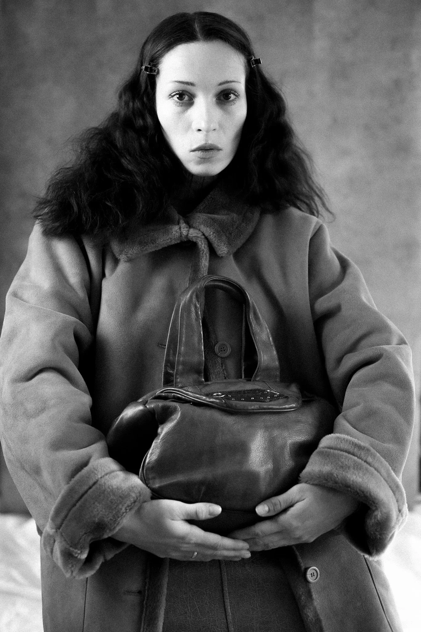 Portrait noir et blanc d'une femme aux cheveux longs et ondulés, portant un manteau et tenant un sac en cuir, avec un fond neutre.