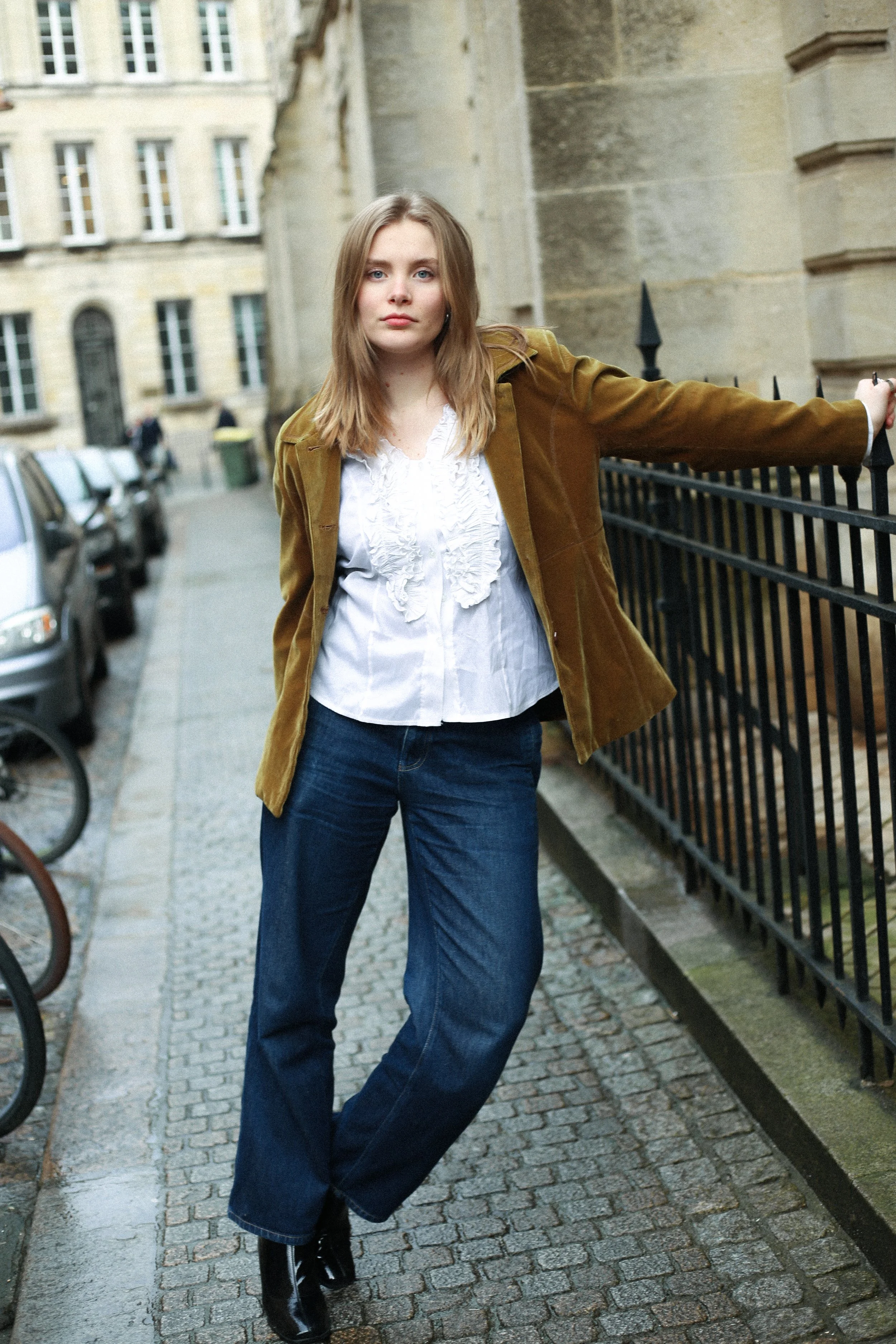 Jeune femme posant sur un trottoir urbain avec bâtiments anciens, porte en arc, voitures en stationnement, vêtements décontractés, un manteau marron, chemise blanche et jeans.