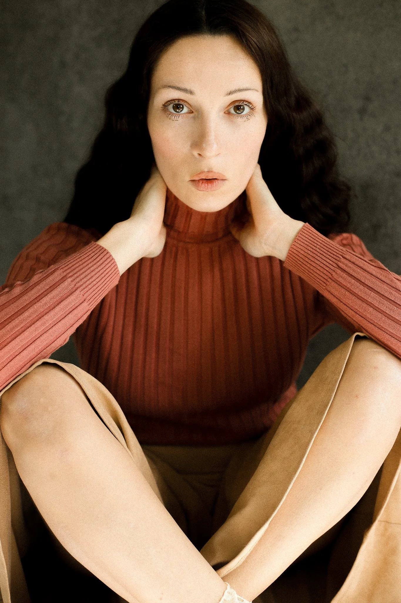 Une jeune femme aux cheveux bruns foncés, portant un pull côtelé rouge et un pantalon beige, assise par terre contre un mur sombre, regardant directement l'appareil photo. Photographe Bordeaux de mode. Editorial. Shooting. Stylisme à Bordeaux.