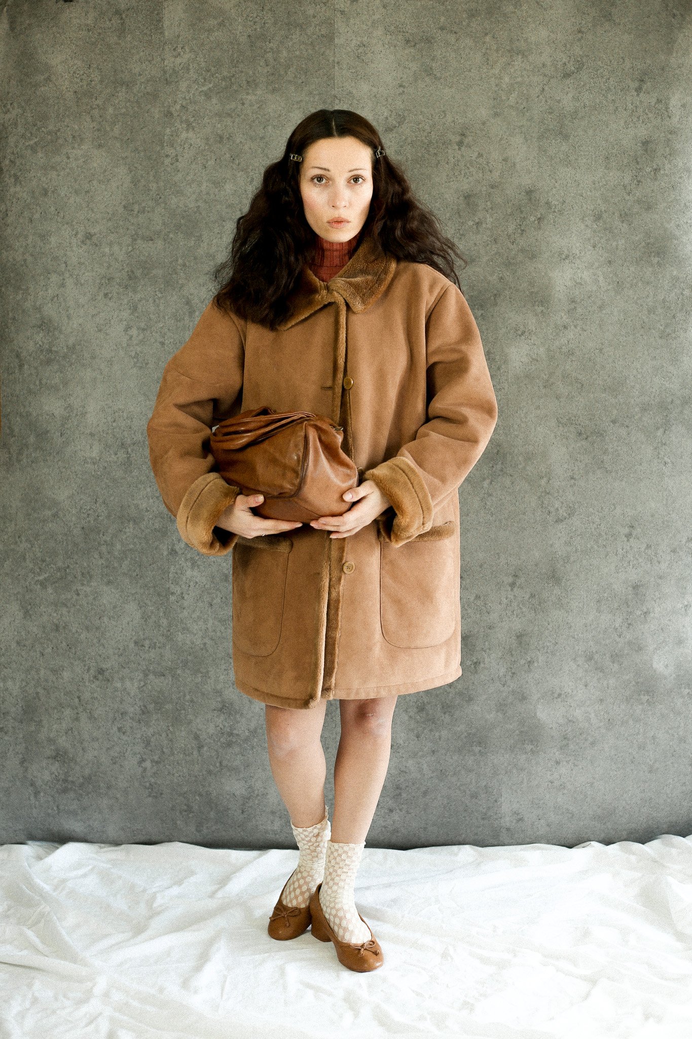 Une femme avec des cheveux longs et foncés porte un manteau marron clair, des chaussettes beige à motif, et des chaussures marron. Elle tient un sac en cuir marron, devant un fond gris et au sol un tissu blanc.Photographe Bordeaux de mode. Editorial.