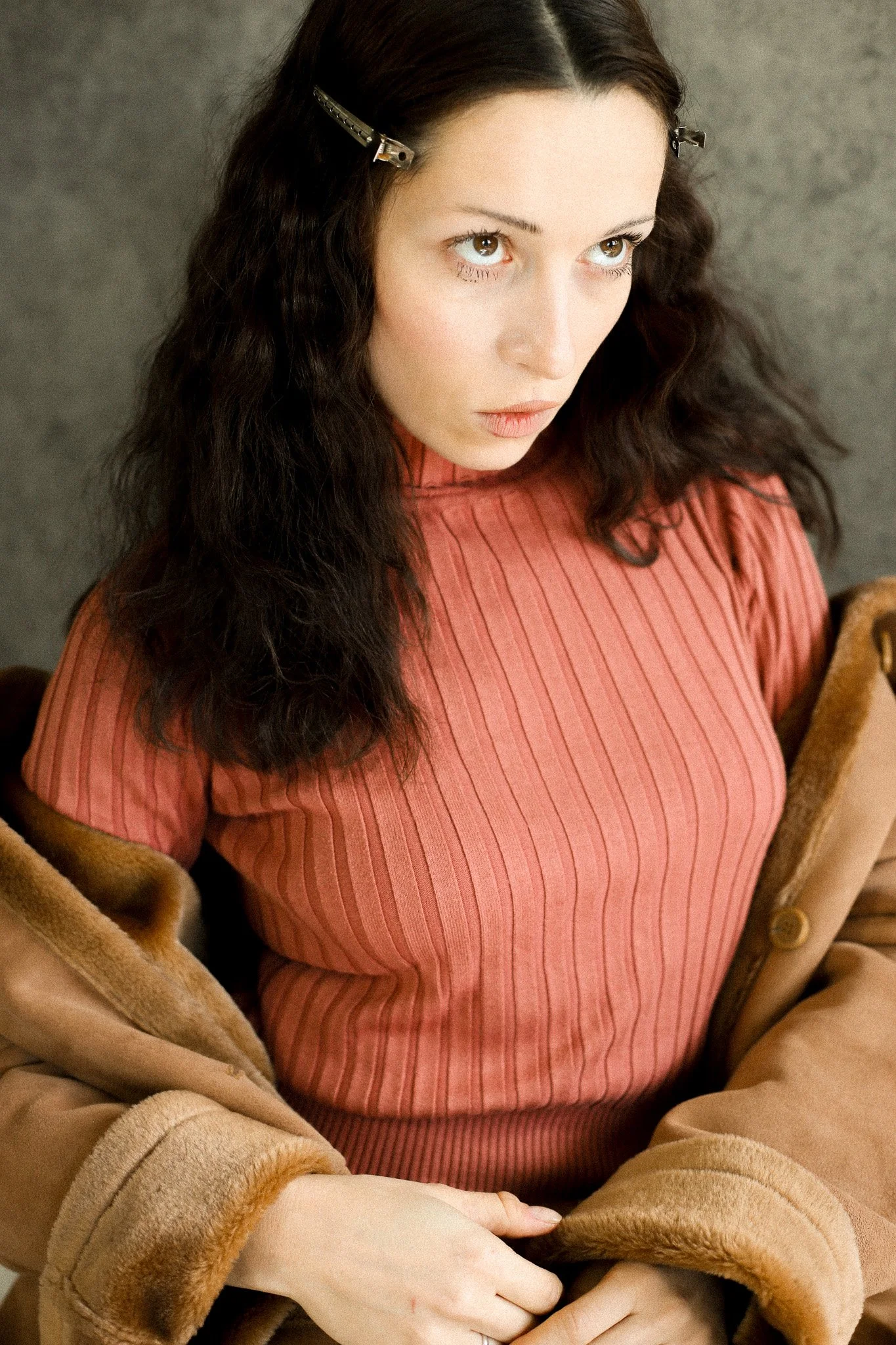 Une femme aux cheveux bruns longs, portant un pull côtelé orange et une veste beige en peluche, assise contre un mur gris.