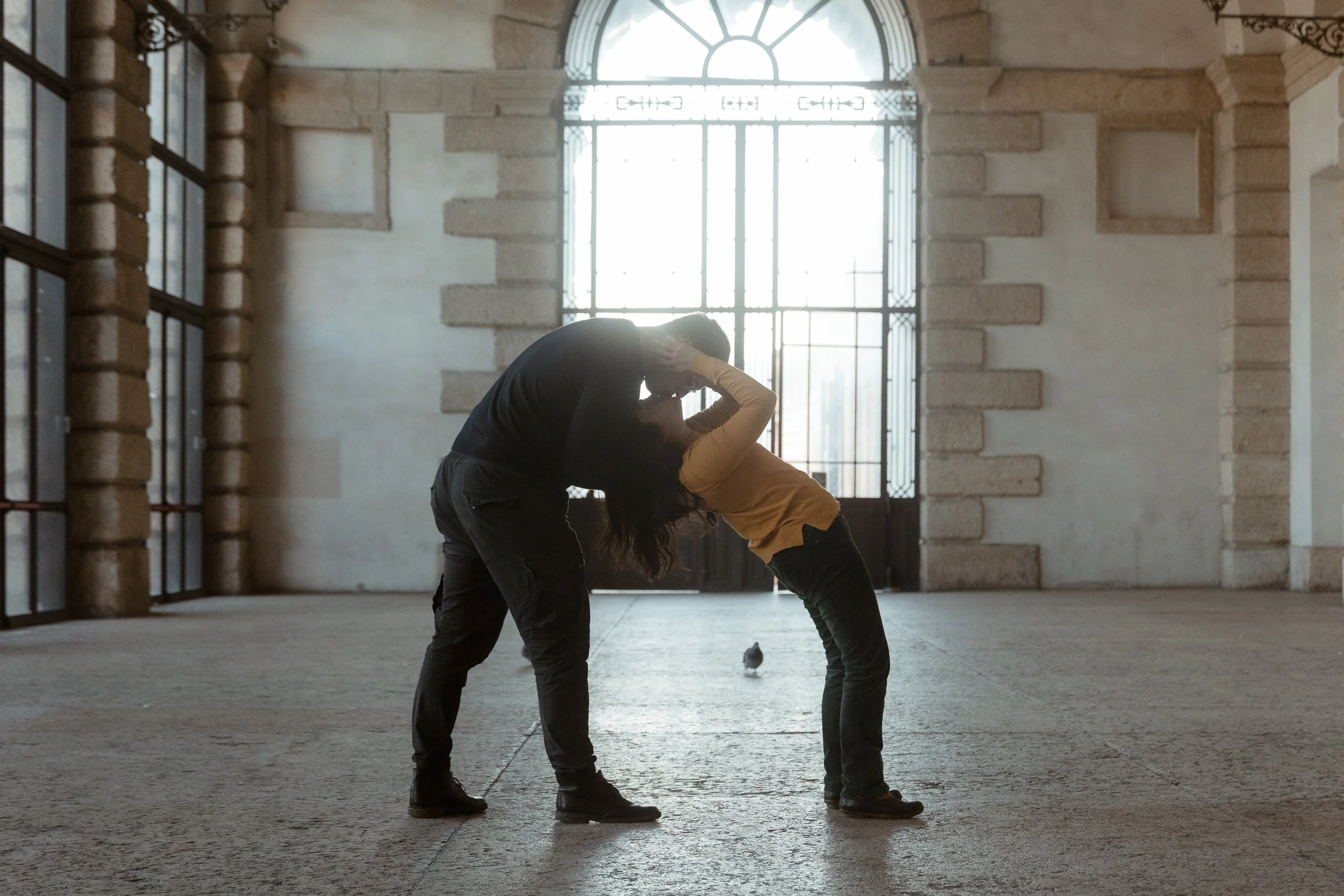 Un uomo e una donna si stanno baciando all'interno di un ambiente spazioso con grandi finestre e pareti di pietra.