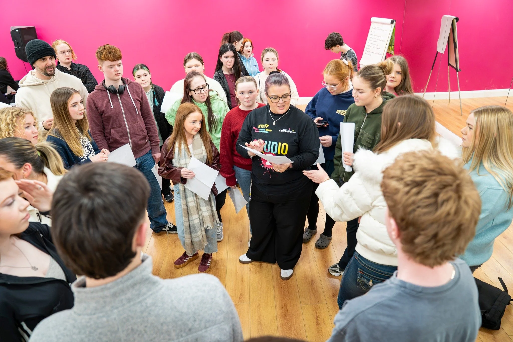 A group of children and adults gathered around a woman with glasses and a black sweatshirt with the word 'STUDIO' on it, in a room with pink walls and a whiteboard. The woman is holding papers and appears to be instructing or leading a discussion.