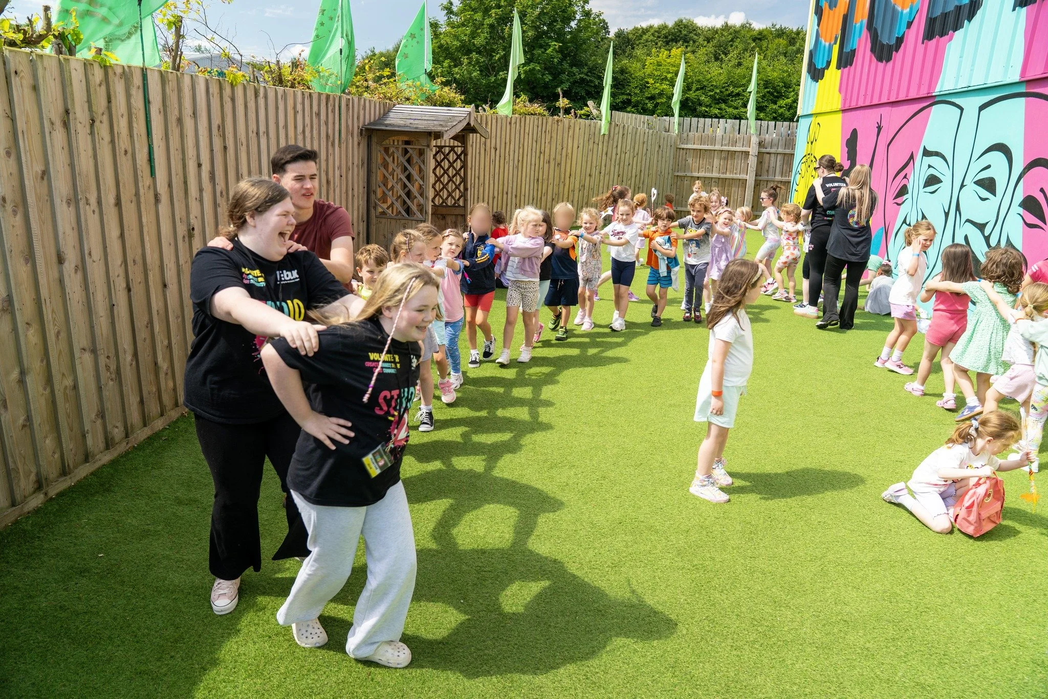 Group of children and volunteers participating in an outdoor activity in a fenced yard with a colorful mural wall. The children are line dancing or playing a game, while the volunteers guide them.
