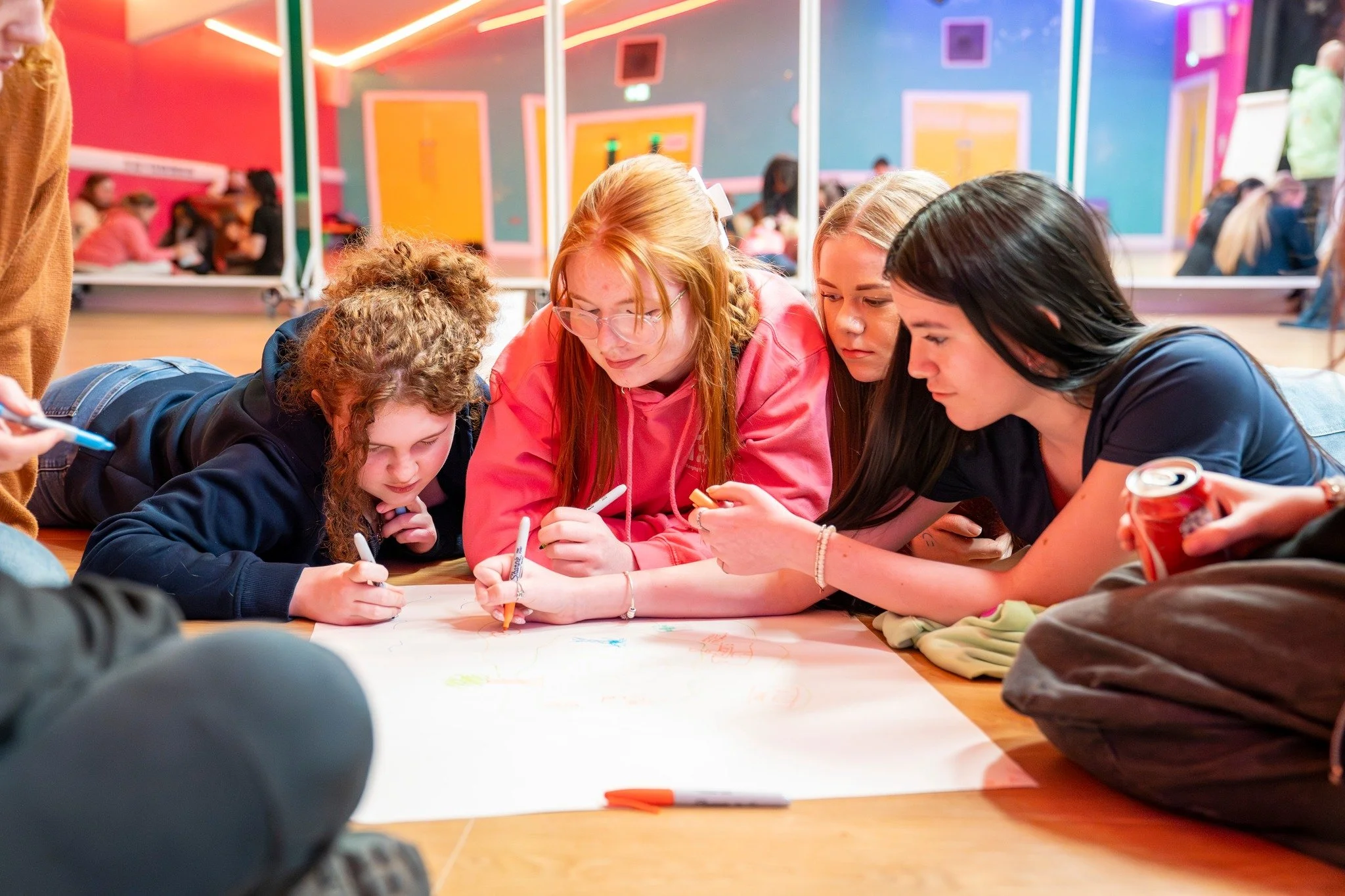 A group of young people, including three girls, lying on the floor together, drawing on large sheets of paper with markers inside a colorful indoor space.
