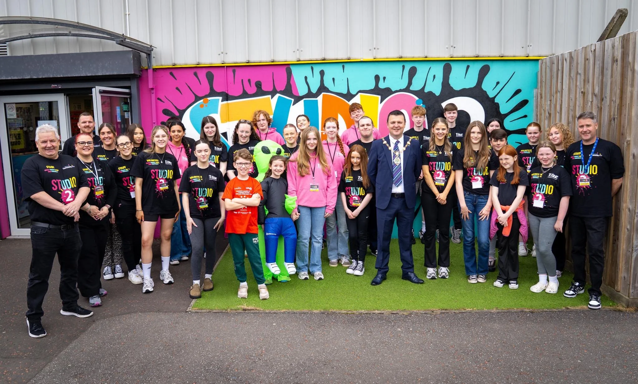 Group of children and adults standing outside in front of a colorful wall with graffiti that says 'STUDIO 2'. They are smiling and wearing casual clothes, some with volunteer badges.