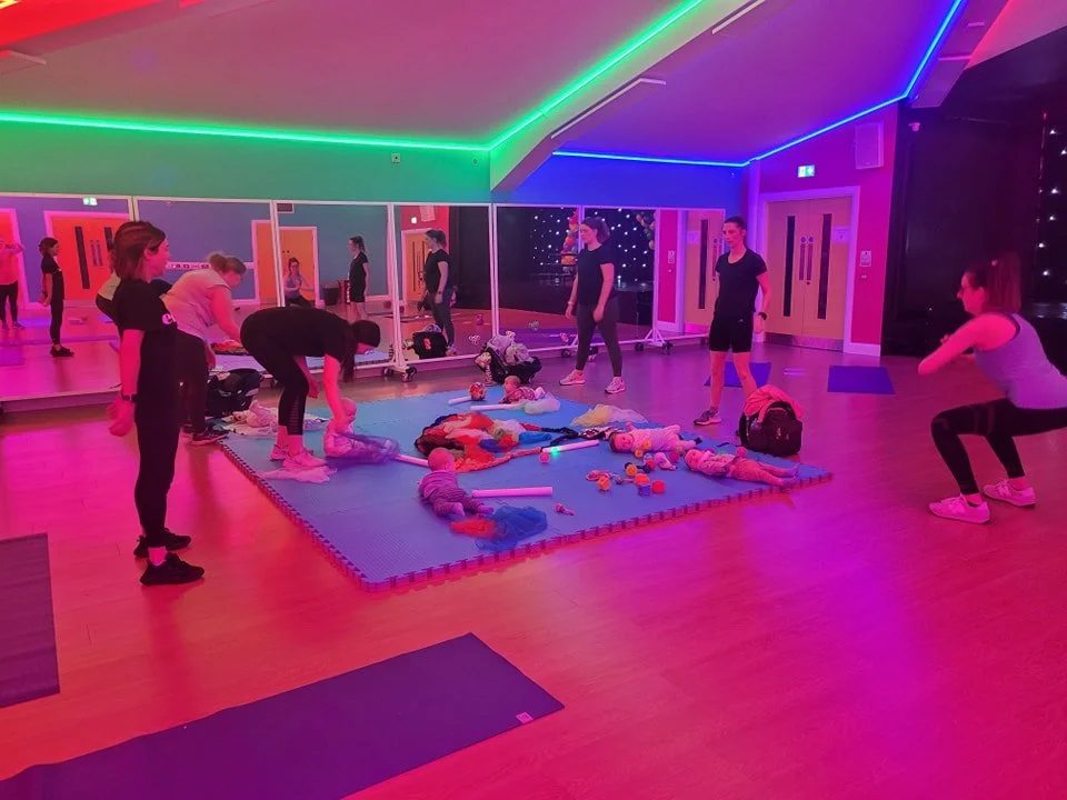 A group of women and babies in a neon-lit fitness or baby yoga class in a room with a pink and purple theme, led by an instructor.