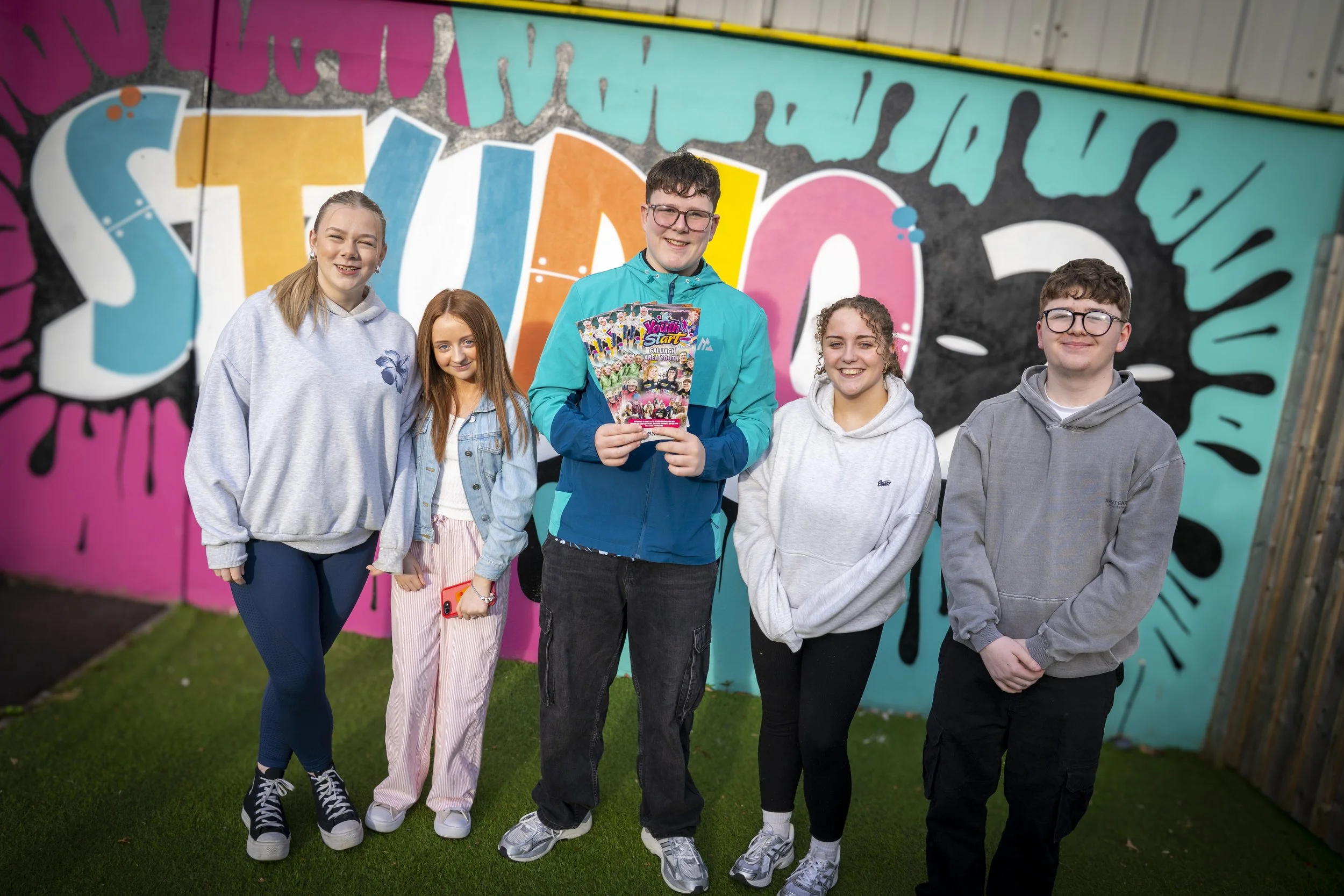Group of five teenagers standing outdoors in front of a colorful graffiti wall, smiling at the camera. One boy in the center is holding a brochure titled 'Youth Start' and wearing a bright blue jacket. Youth Start - Galliagh Area Youth Programme in D