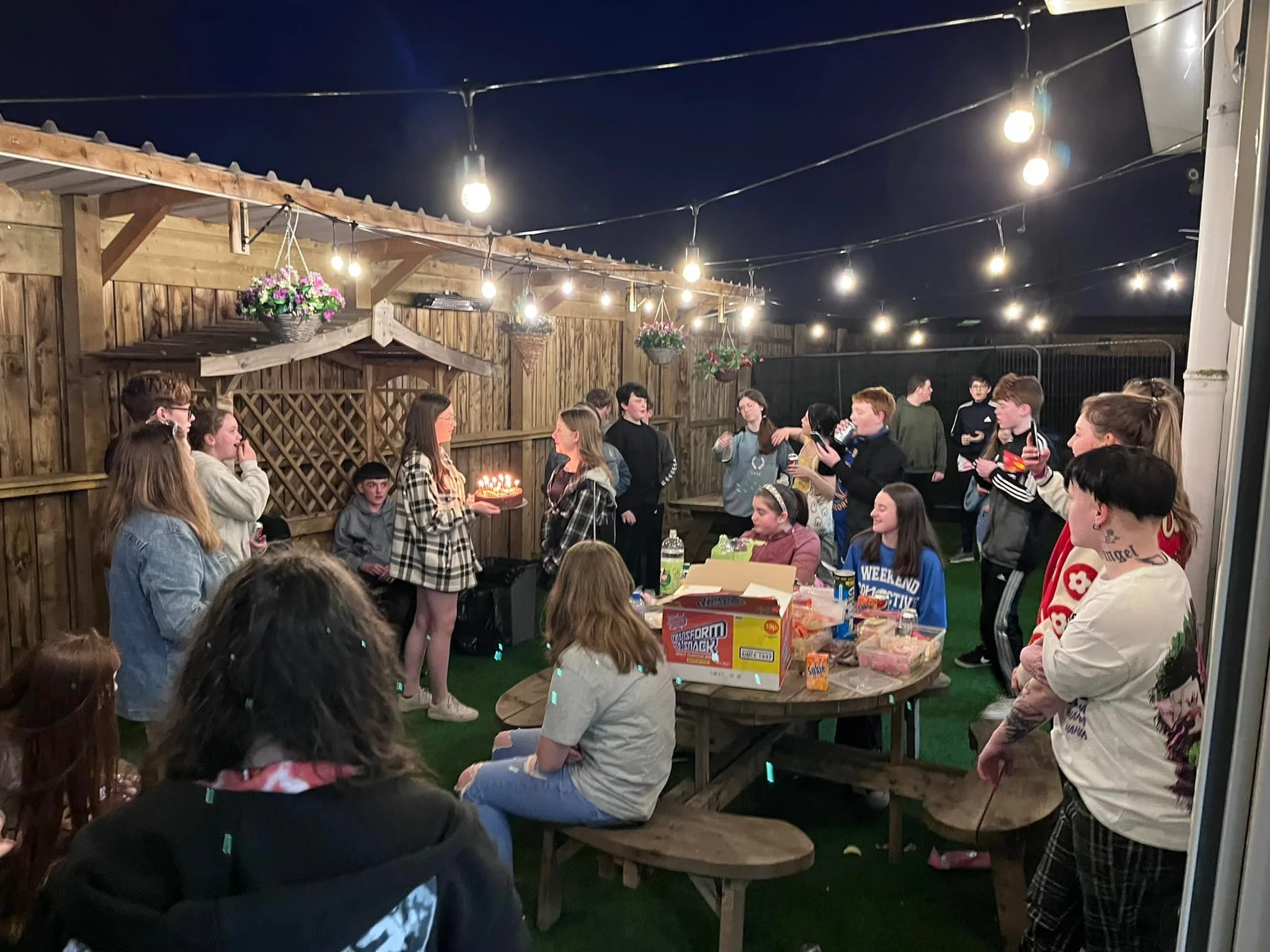People celebrating a birthday party outdoors at night, with a girl holding a cake with lit candles, surrounded by friends and family under string lights, with a wooden fence and hanging flower baskets in the background.