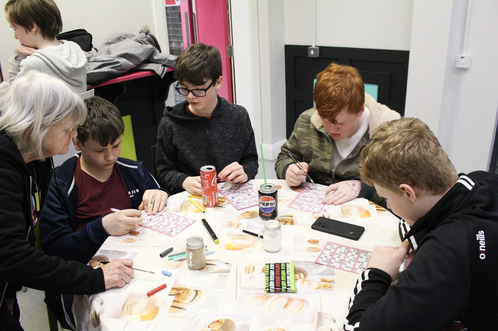 A group of six people, five young men and one older woman, sitting around a table covered with bingo cards, markers, drinks, and snacks, engaged in a bingo game in an indoor setting.