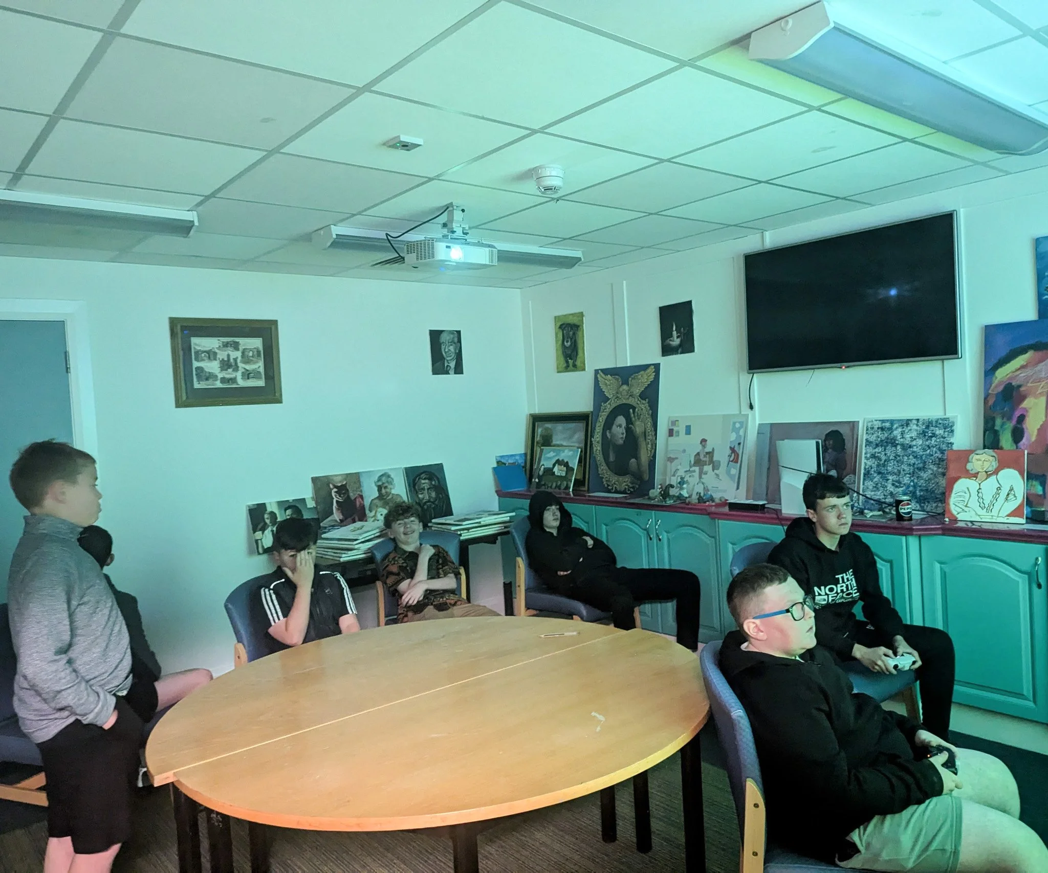 Group of young boys sitting and standing in a room with artwork and a large television on the wall, some holding video game controllers.