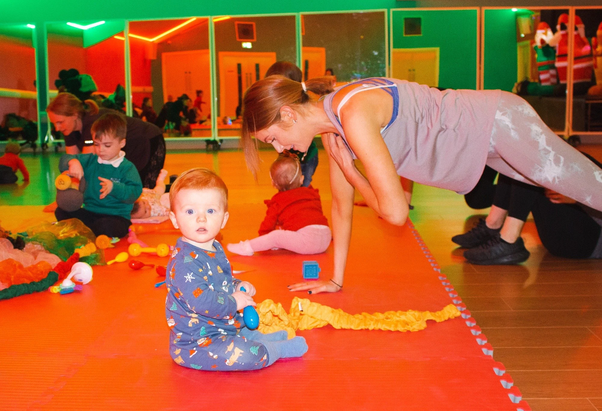 A group of children and adults in an indoor play area with colorful lighting. A young girl with red hair and blue eyes, wearing pajamas with playful prints, is sitting on an orange mat holding a toy. Nearby, a woman with light brown hair is in a plan