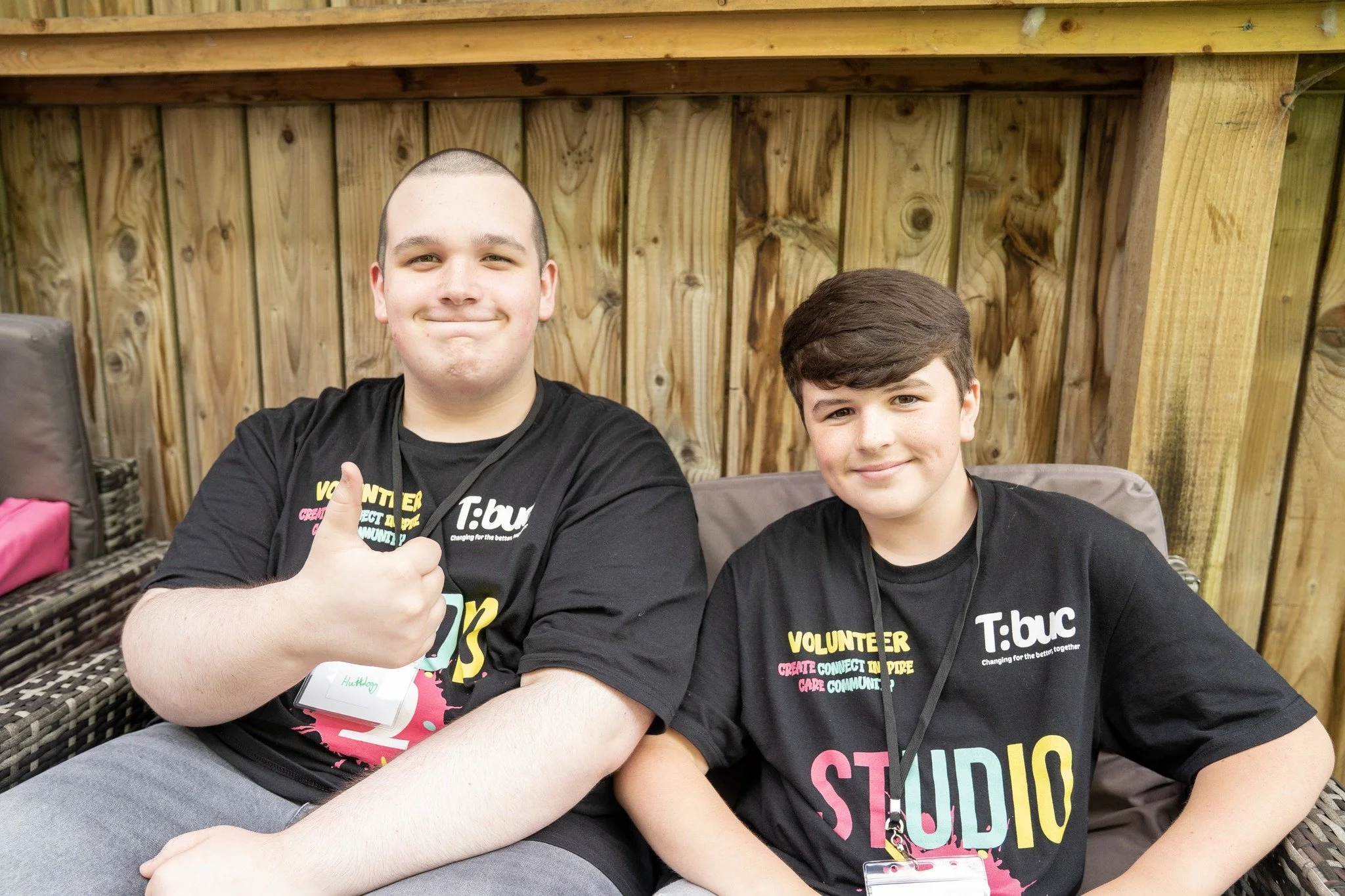 Two young males sitting outdoors in front of a wooden fence, wearing black T-shirts with colorful text about volunteering. The male on the left has a buzz cut and is smiling with his eyes slightly squinted, while the male on the right has dark hair a