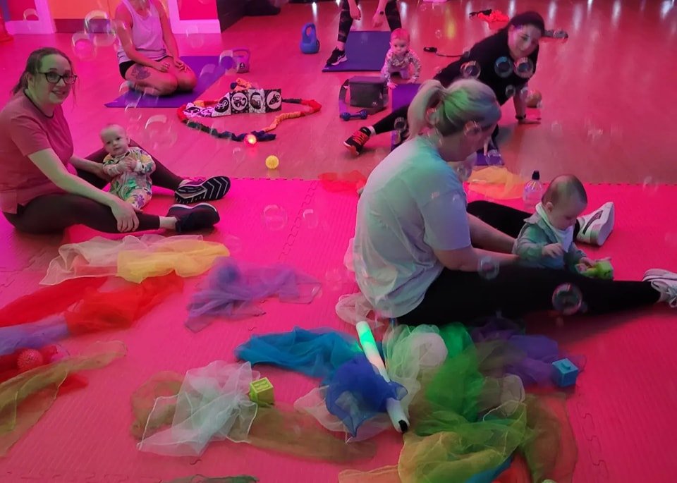 Parents and children participating in a colorful indoor play or exercise class on red mats, surrounded by toys, bubbles, and vibrant decorations.