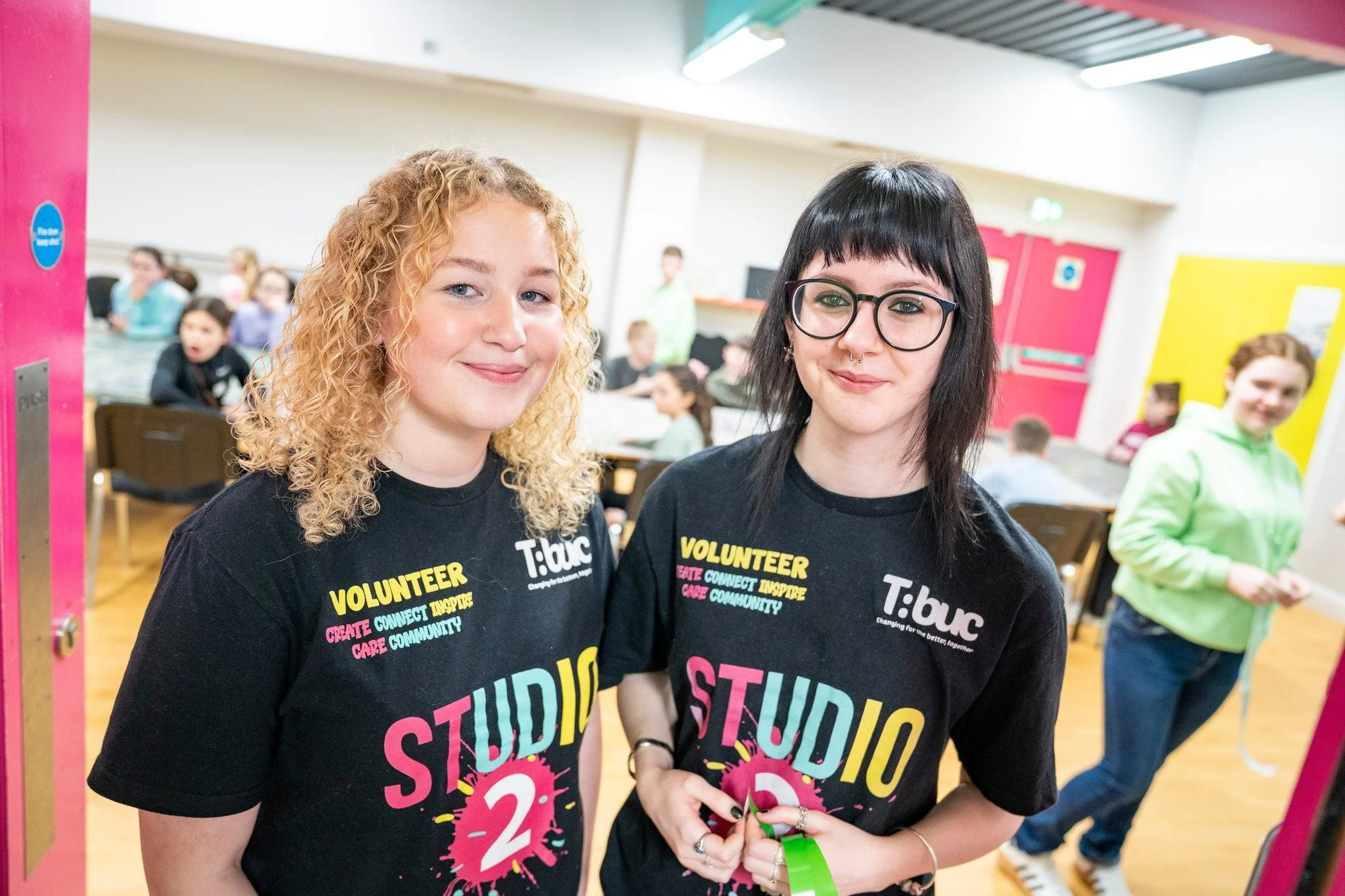 Two young women with curly and straight dark hair wearing black 'Volunteer' t-shirts standing inside a brightly lit community center, smiling at the camera. Several other people are visible in the background.