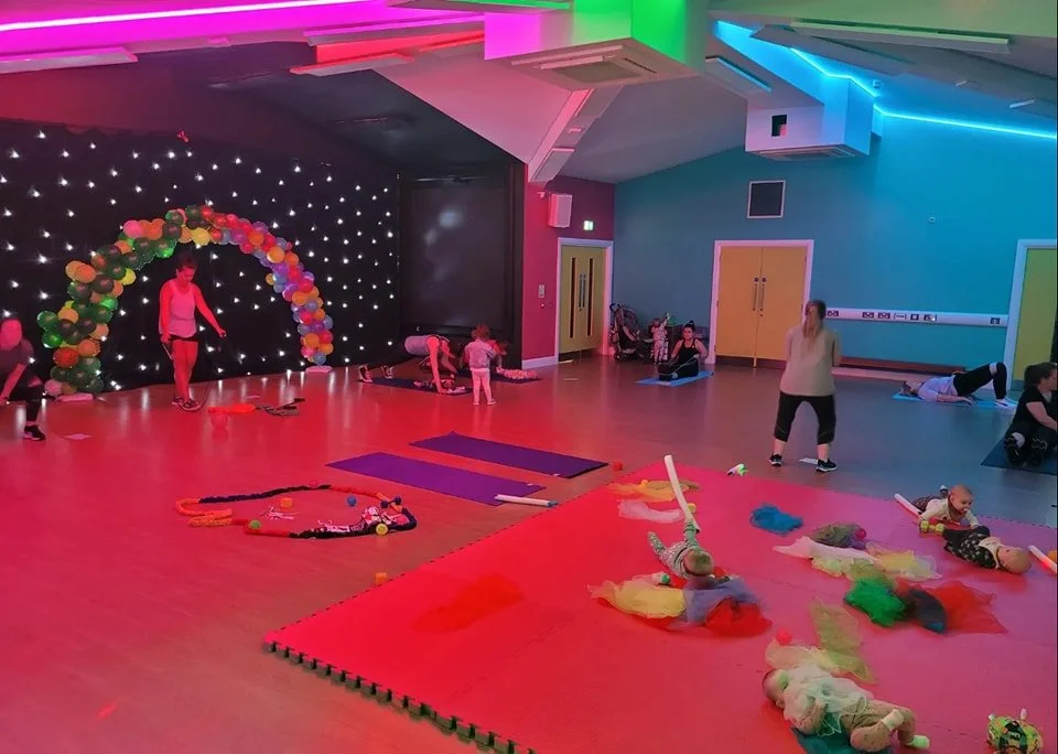 Children and adults participating in a gymnasium class with colorful lighting, balloons, mats, and toys.