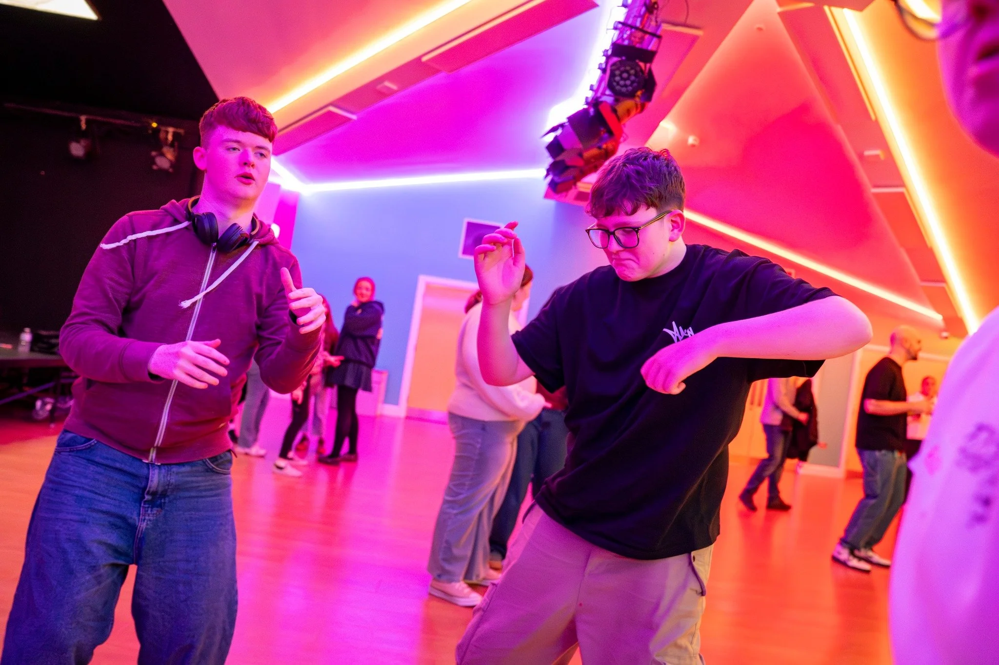 Two young men dancing at a party or nightclub with vibrant pink and purple lighting, surrounded by other people.