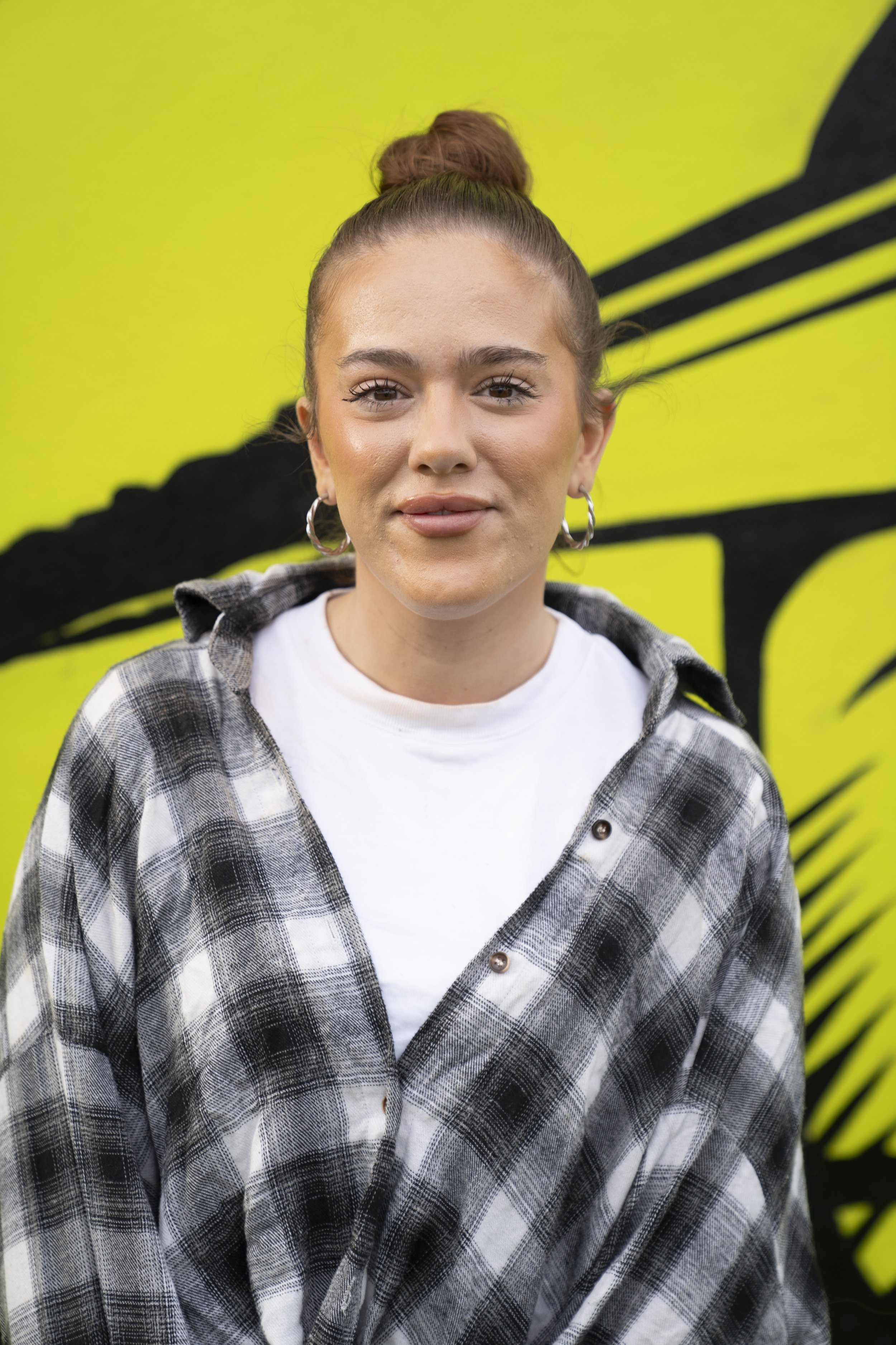 A young woman with her hair in a bun, wearing hoop earrings, a white t-shirt, and a black and white plaid shirt, standing in front of a yellow background with black graphic design.