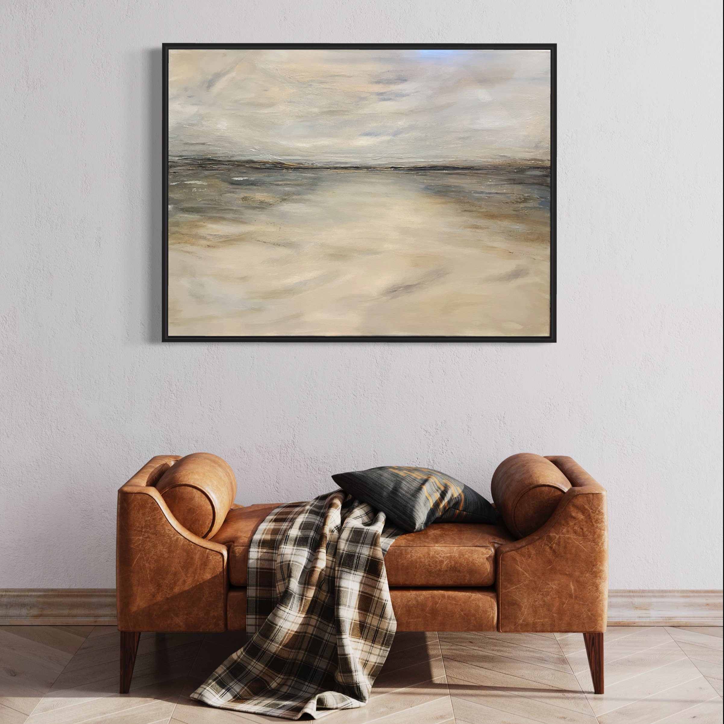 A vintage brown leather sofa with a checkered blanket and a pillow, set against a white wall with a large abstract painting hanging above it.