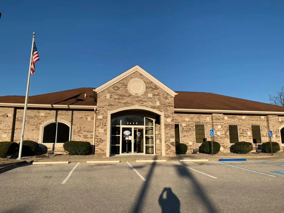 A brick commercial building with a glass entrance and the address 4451 above the door. The building has several windows and bushes in front. There are parking spaces, including handicap spots, directly in front of the building, with an American flag on a flagpole nearby against a clear blue sky.