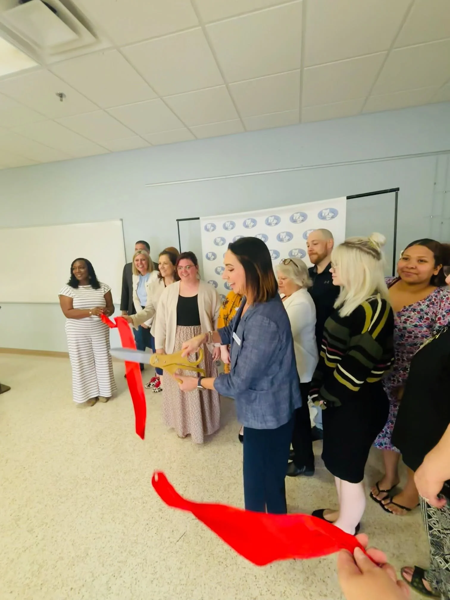 Group of diverse people standing in a line for a ceremony or event in a room with a blue wall and a whiteboard. Someone is holding a red ribbon at the front.