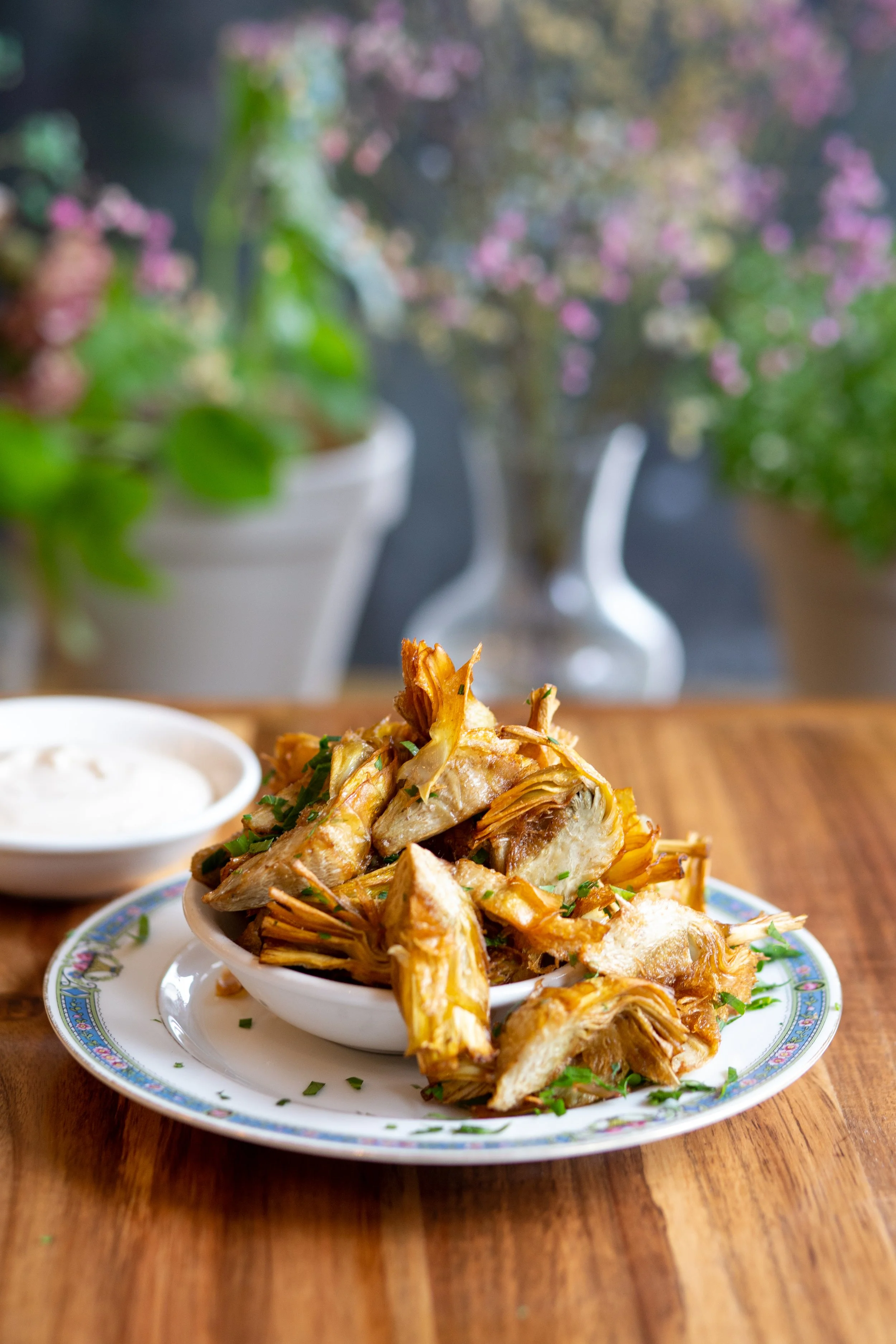 A plate of fried artichoke hearts garnished with chopped herbs, with a side of dipping sauce on a wooden table, and potted plants and flowers in the background.
