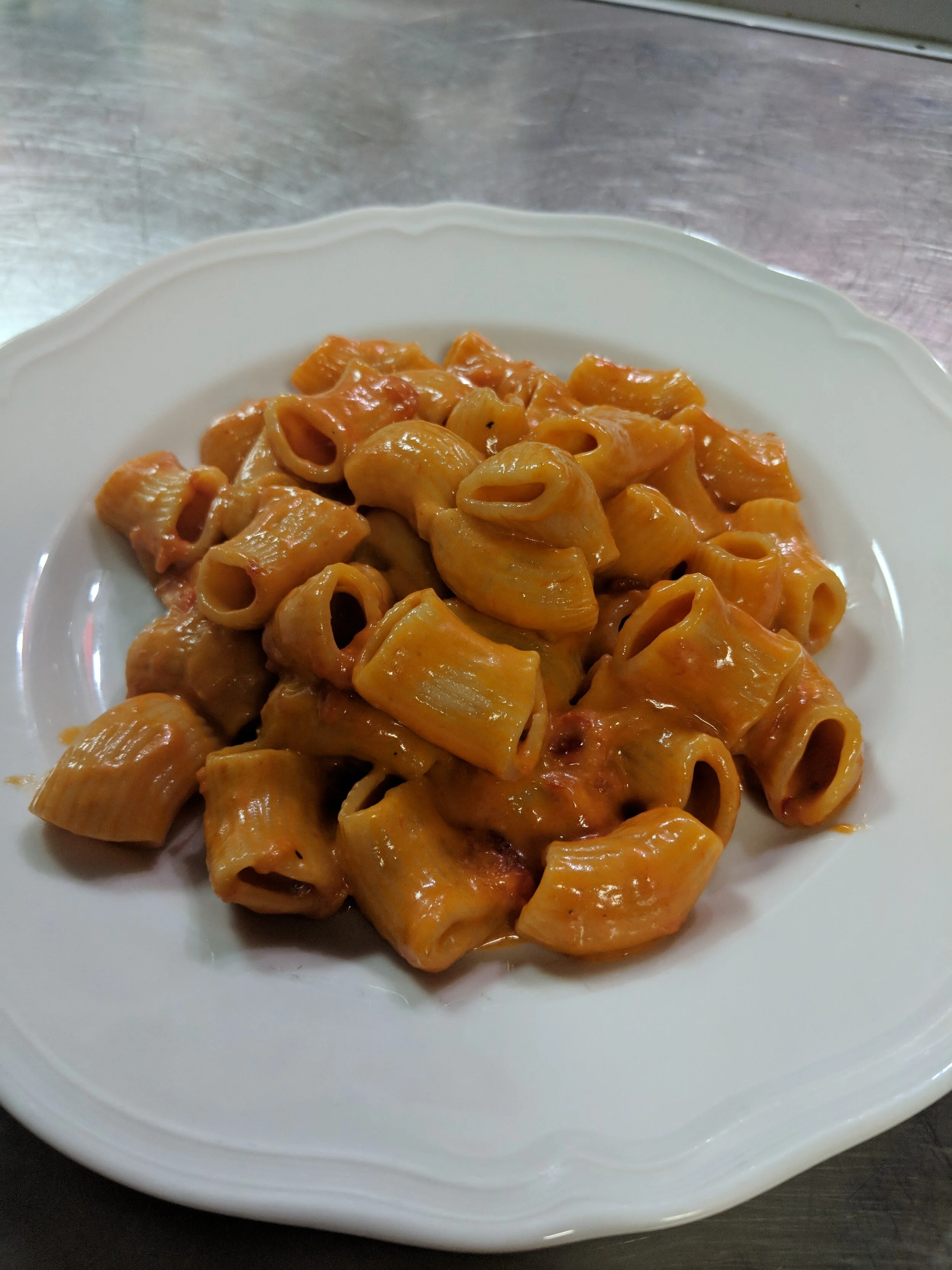 A white plate of cooked pasta with a tomato-based sauce on a stainless steel surface.