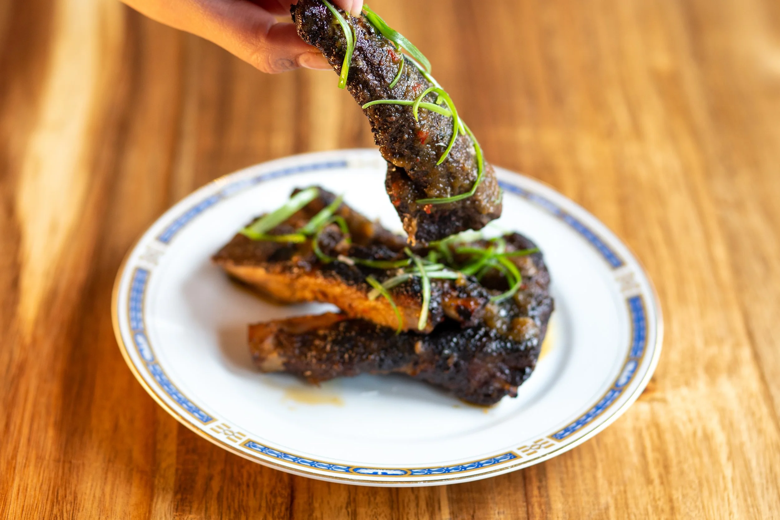 Close-up of grilled beef short ribs garnished with chopped green onions on a decorative white plate with blue and gold details, placed on a wooden table.