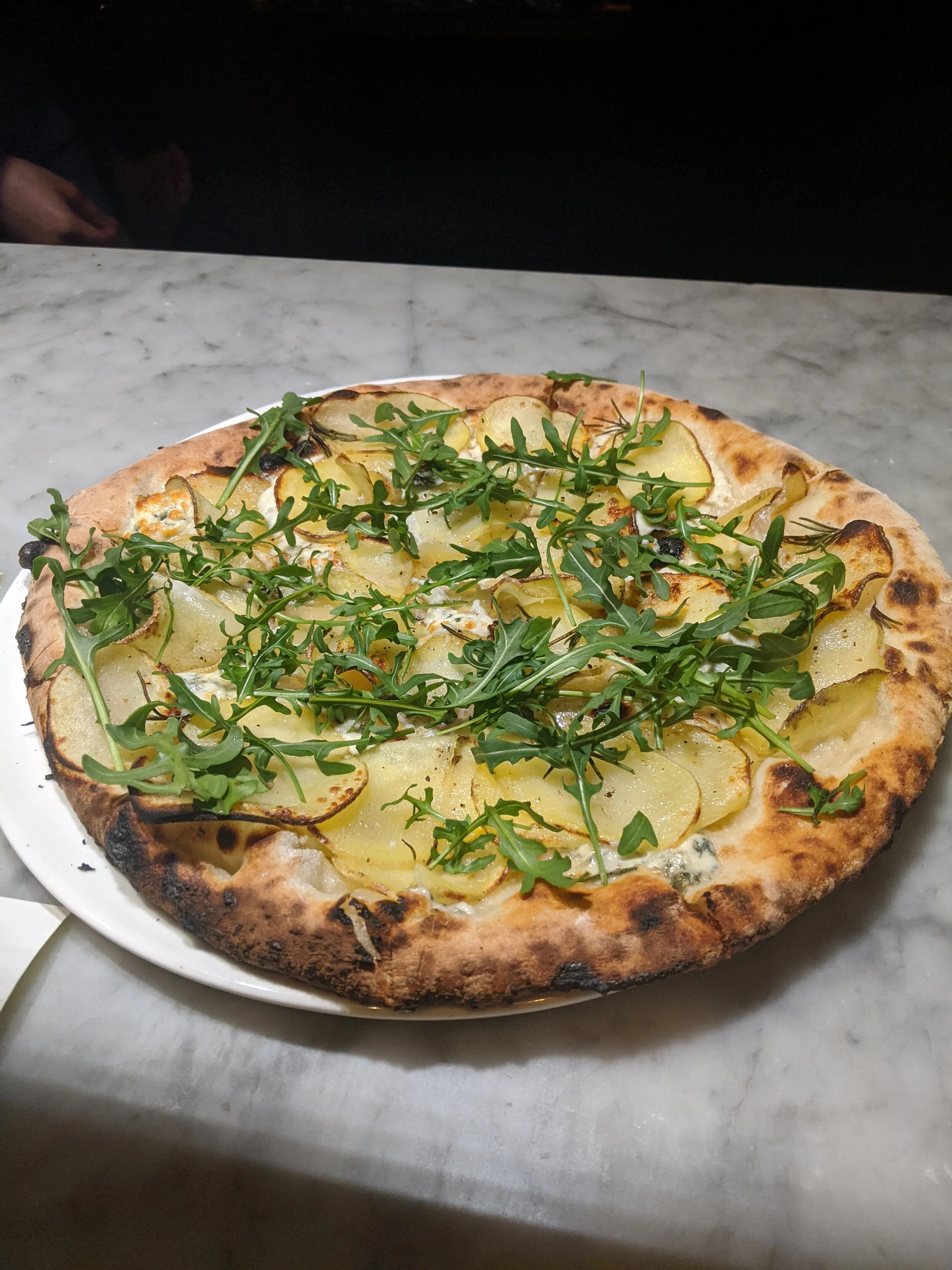 A vegetable pizza topped with arugula leaves, thinly sliced potatoes, and cheese, on a white plate.