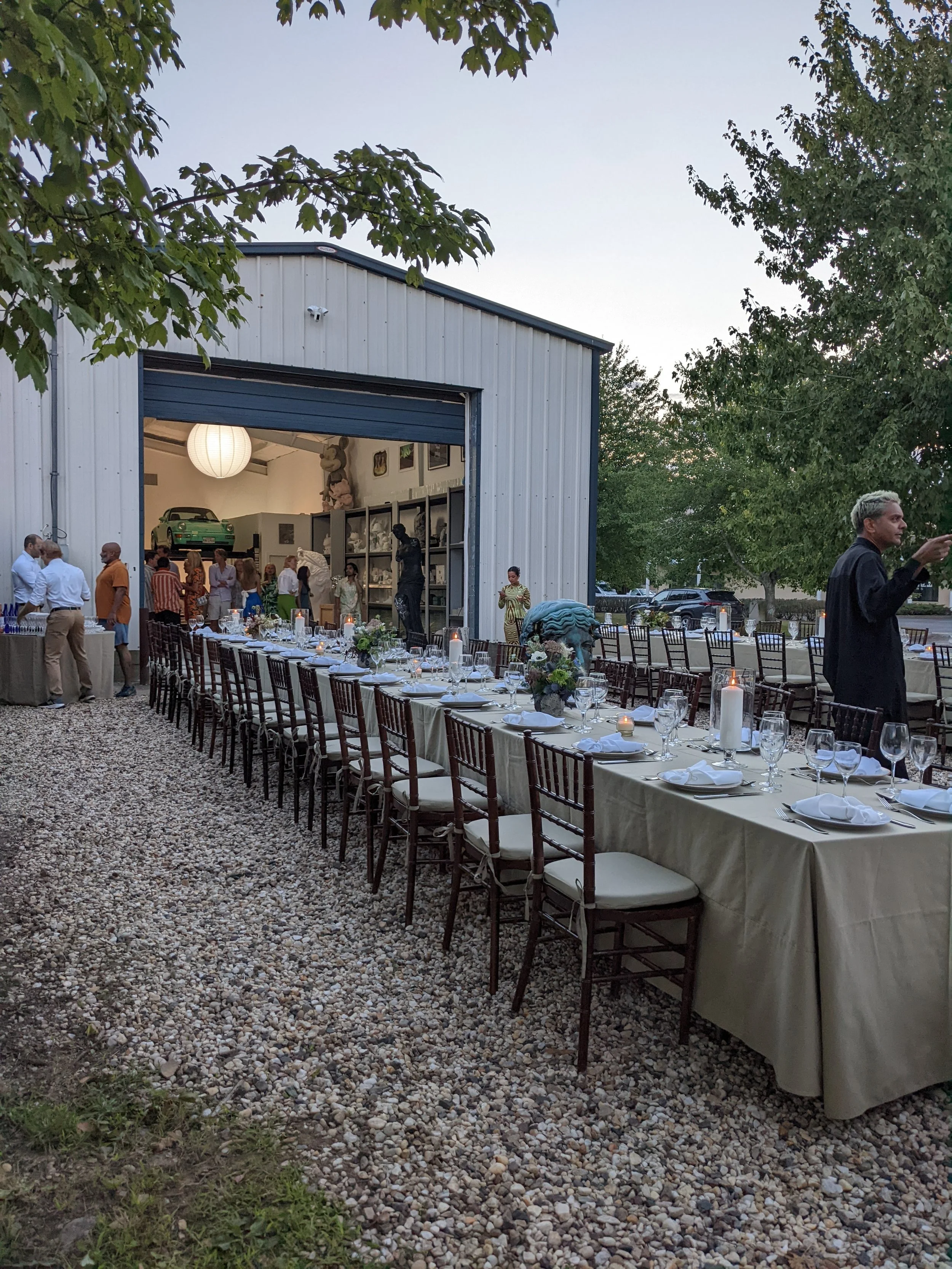 An outdoor event setup with a long dining table featuring candles and floral centerpieces, positioned outside a large open shed with people mingling inside. There are trees surrounding the area, and the scene appears to be during sunset or early even