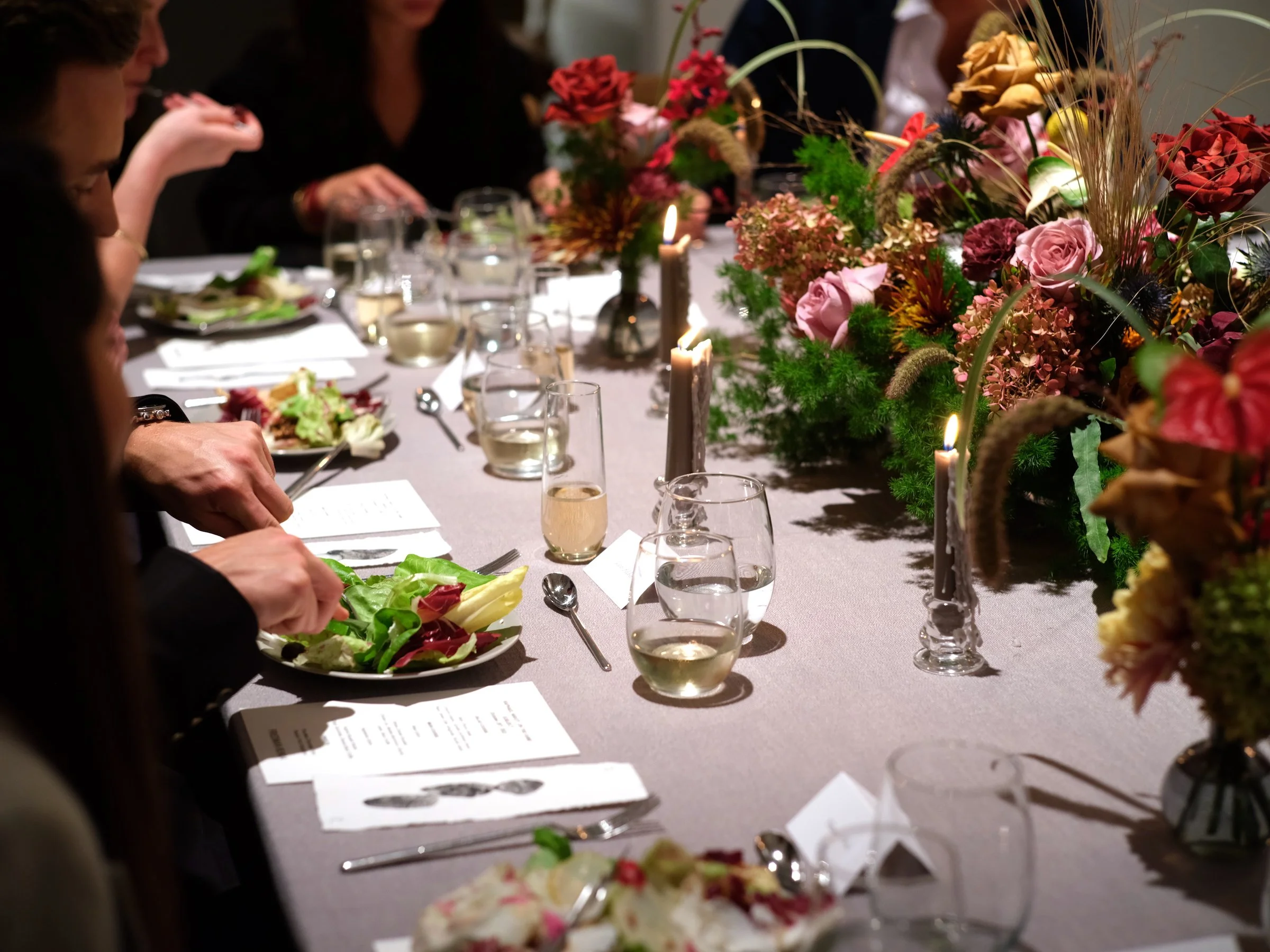 A formal dinner party with a long table decorated with flowers, candles, and place settings including salad plates, wine glasses, and menus.