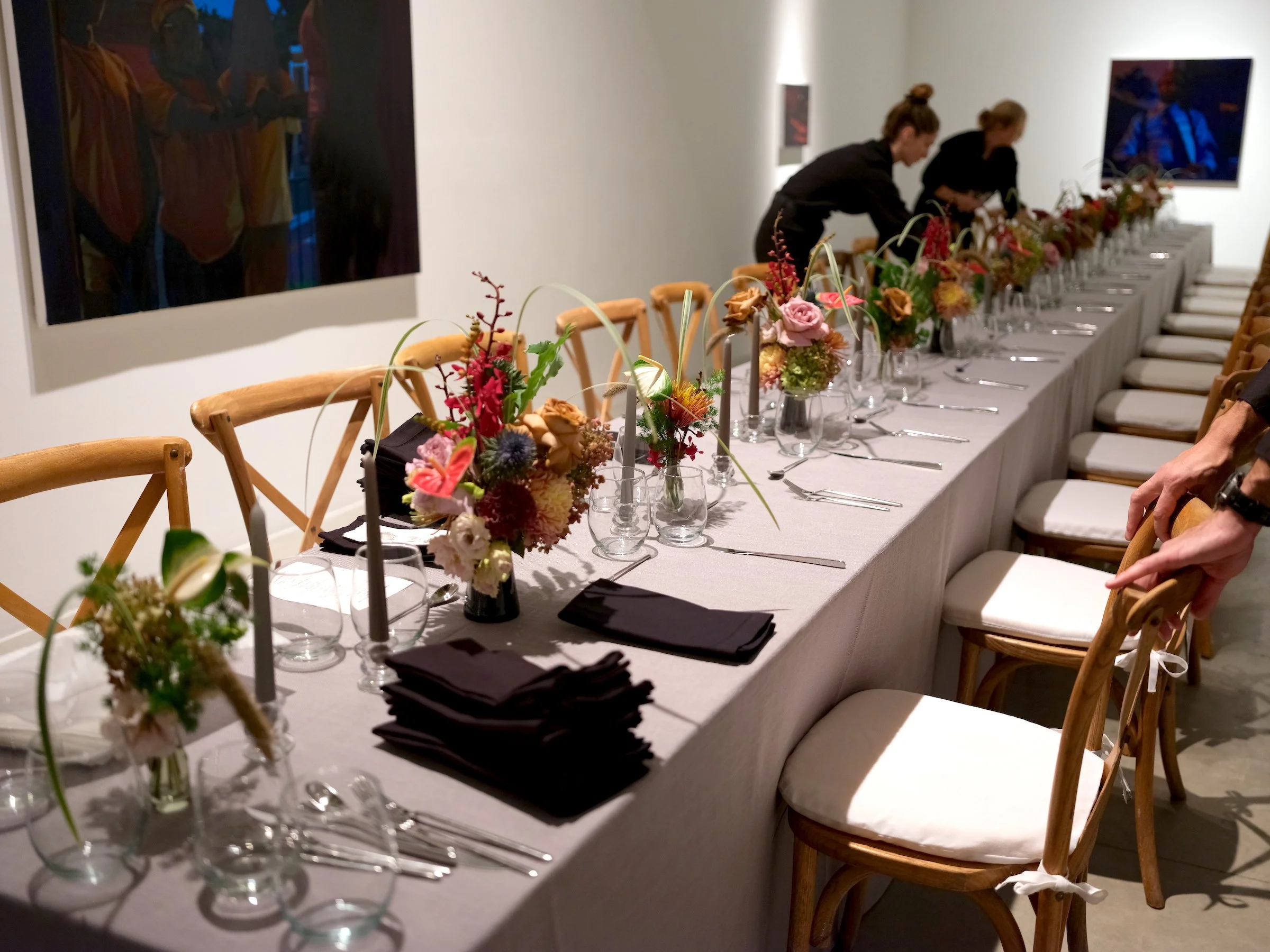 Long dining table set with colorful flower centerpieces, black napkins, glassware, and silverware in an indoor art gallery or event space with people arranging the table.