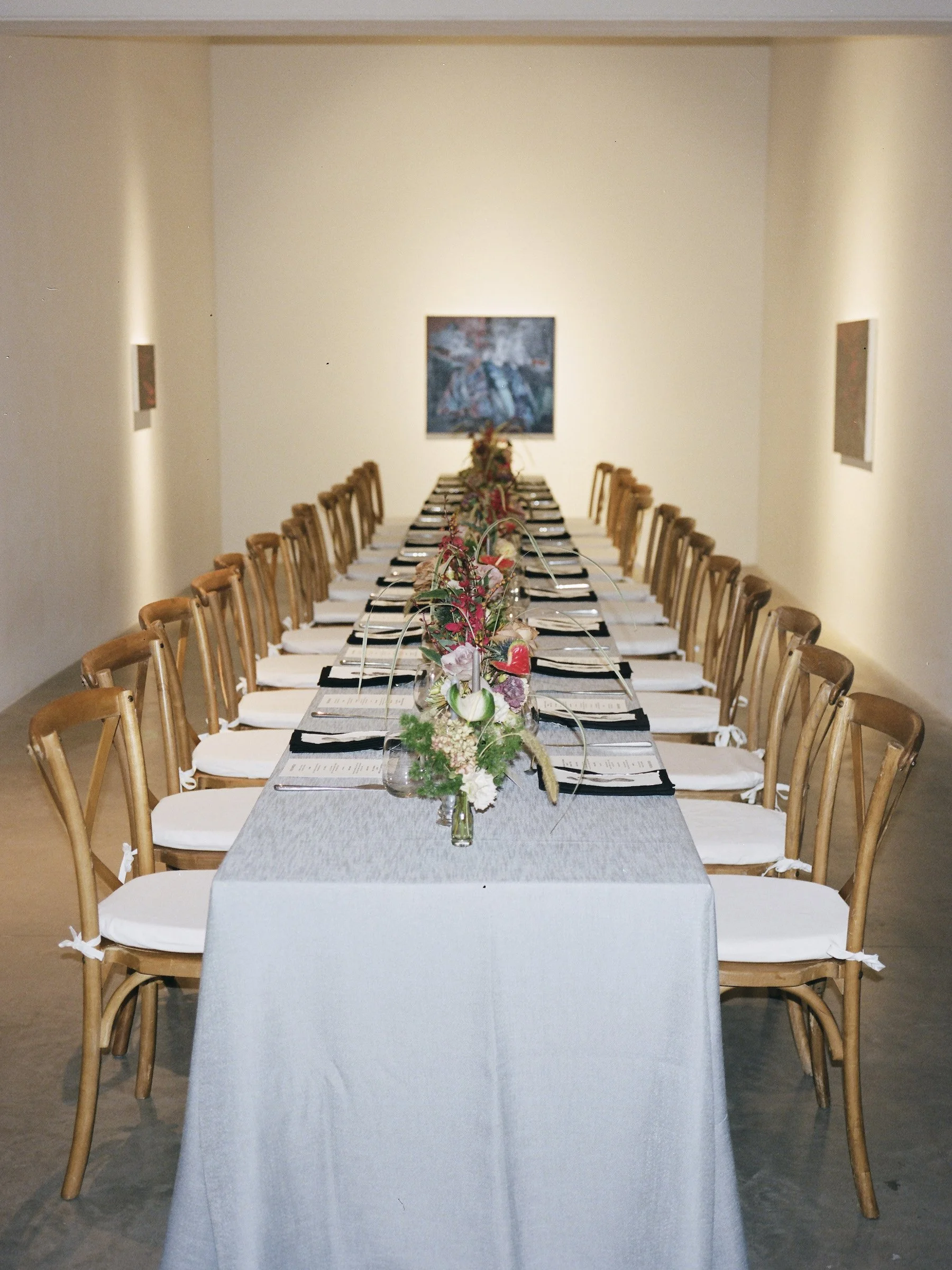 A long dining table set for a special occasion with floral centerpieces, surrounded by wooden chairs with white cushions, in an art gallery or event space with artwork on the walls.