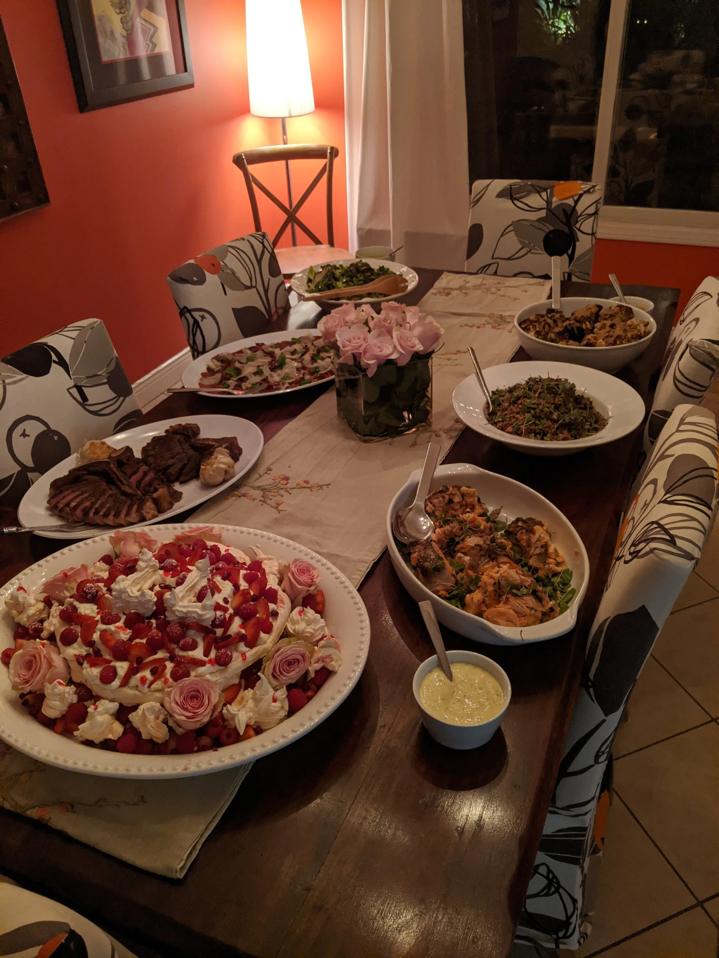 A dining table set with various dishes including a large fruit-topped cake, salads, grilled meats, and other cooked entrees, with chairs around it and a flower centerpiece, in a warmly lit dining room.