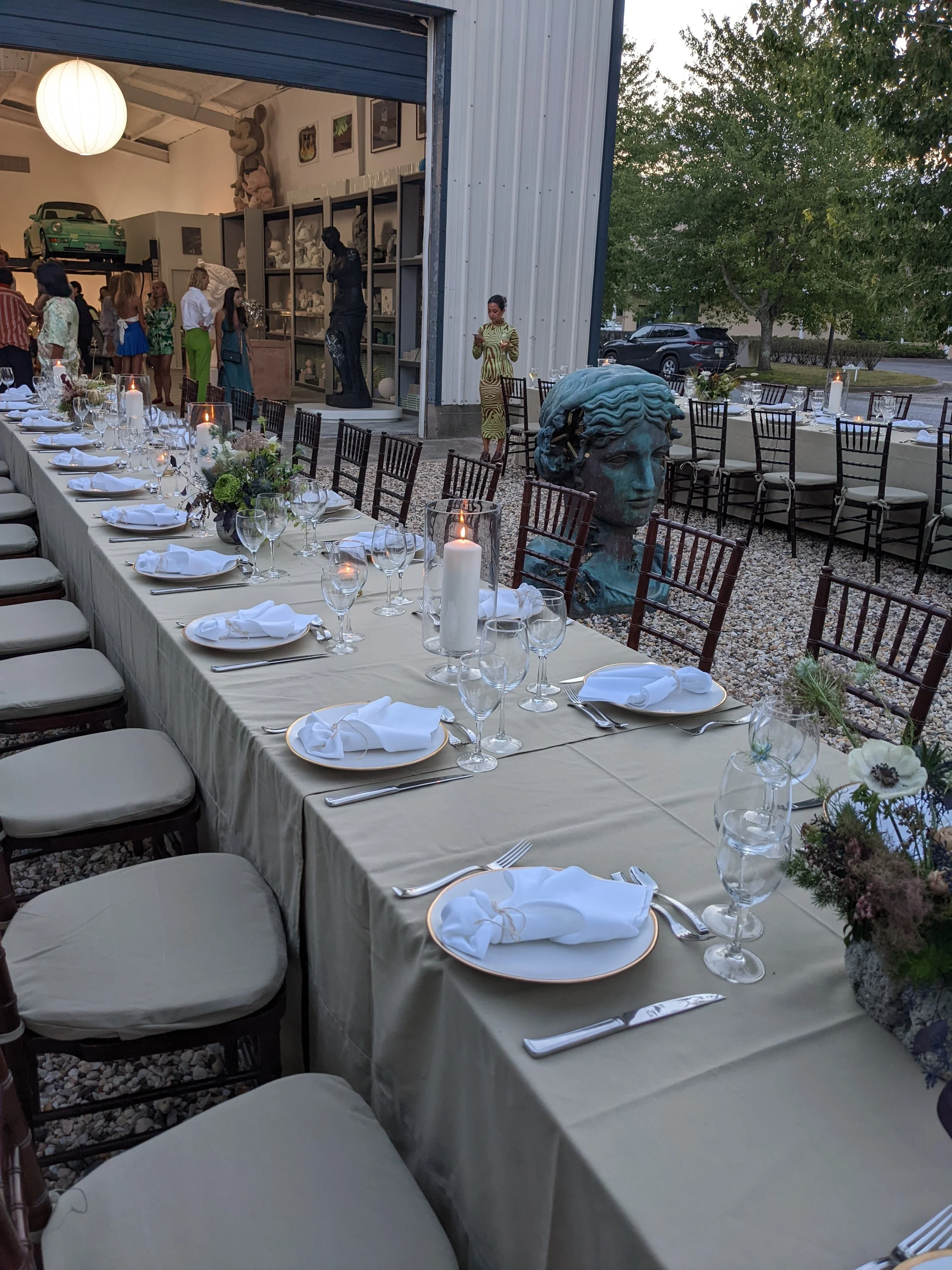 Long banquet table set for an outdoor event with white tablecloths, white napkins, glassware, and candles, next to decorative sculptures in a partially open building with people inside and outside.