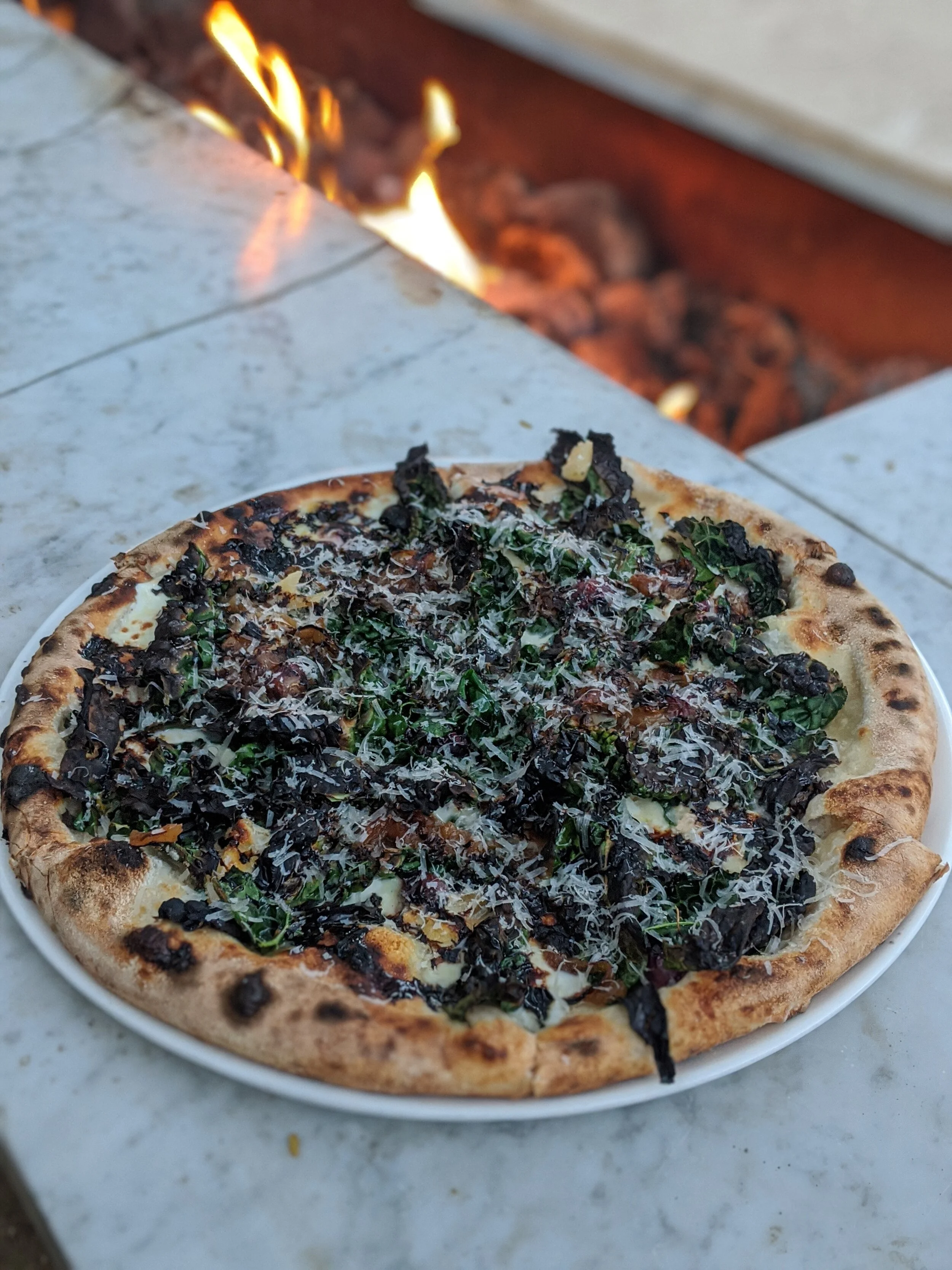 Pizza topped with kale and grated cheese on a white plate, with a fire pit in the background