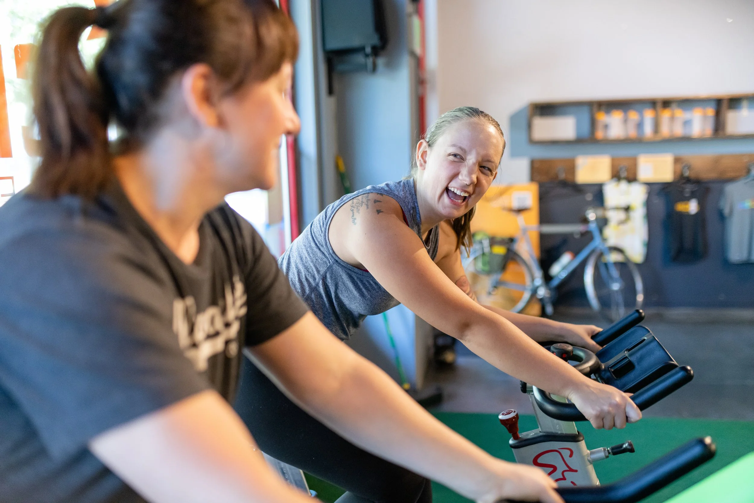 Two women smiling and exercising on stationary bikes in a gym, with one woman in focus and the other blurred in the foreground.