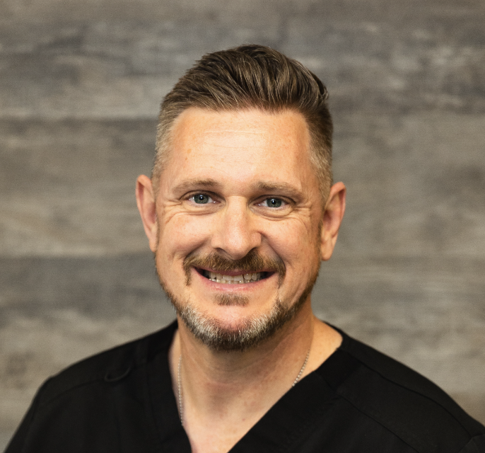 Portrait of a smiling man with short gray hair and a beard, wearing a black shirt, standing in front of a gray wooden wall.