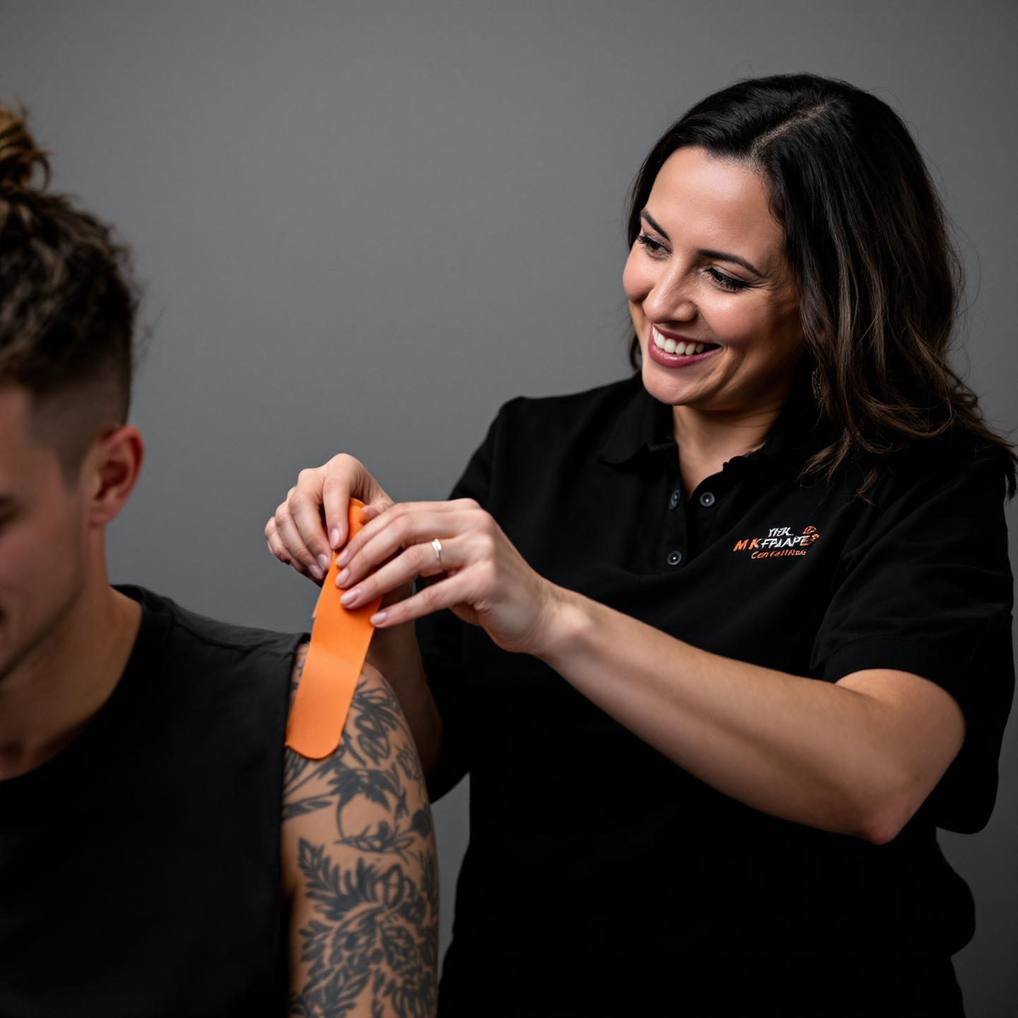 A woman is applying adhesive kinesiology tape to a young man's shoulder, smiling while working in a clinical setting.