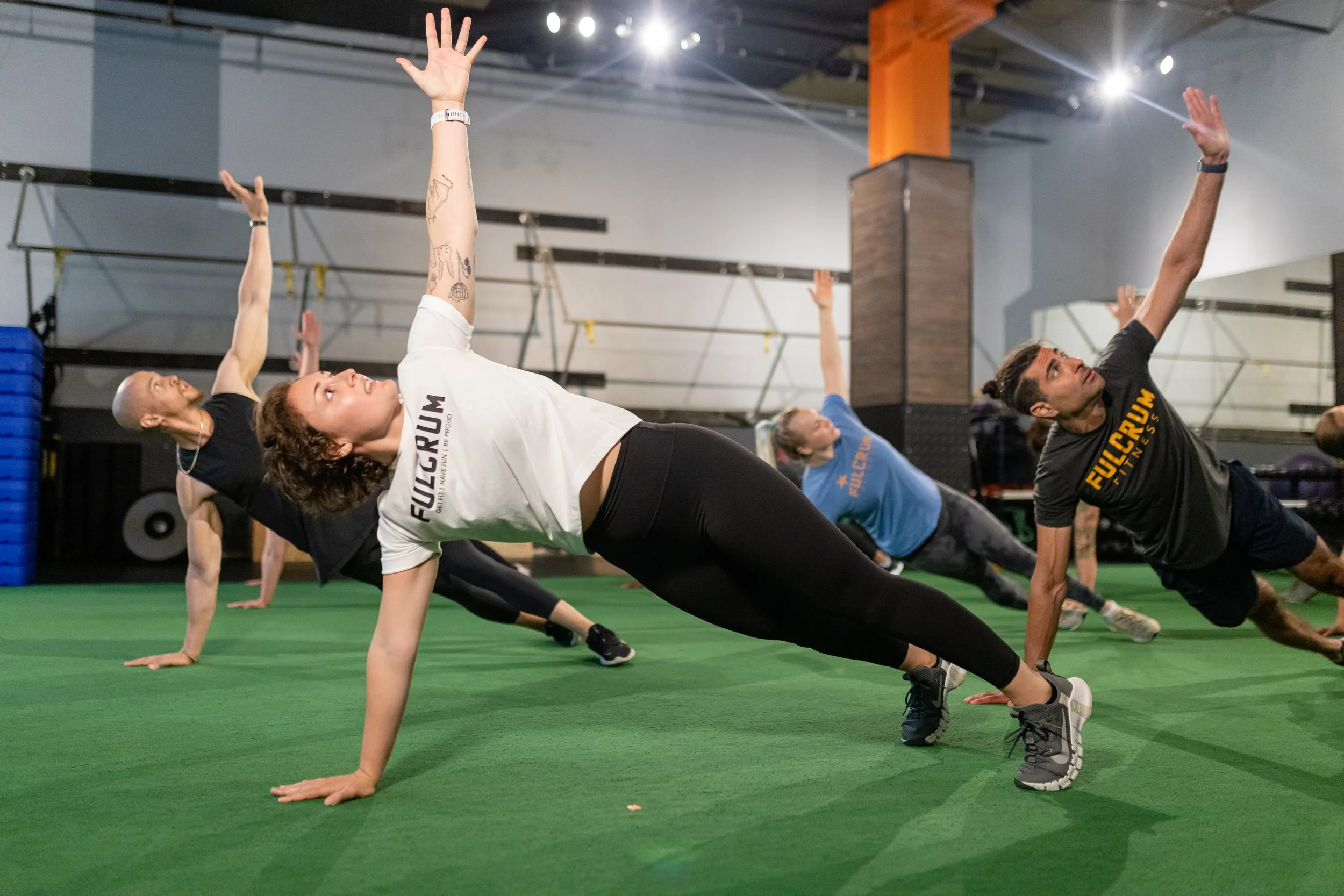 People participating in a fitness class doing low plank side stretches on green turf.