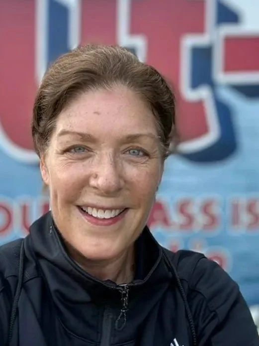 Close-up of a smiling woman with light skin, blue eyes, and brown hair tied back, standing outdoors with a large colorful sign in the background.